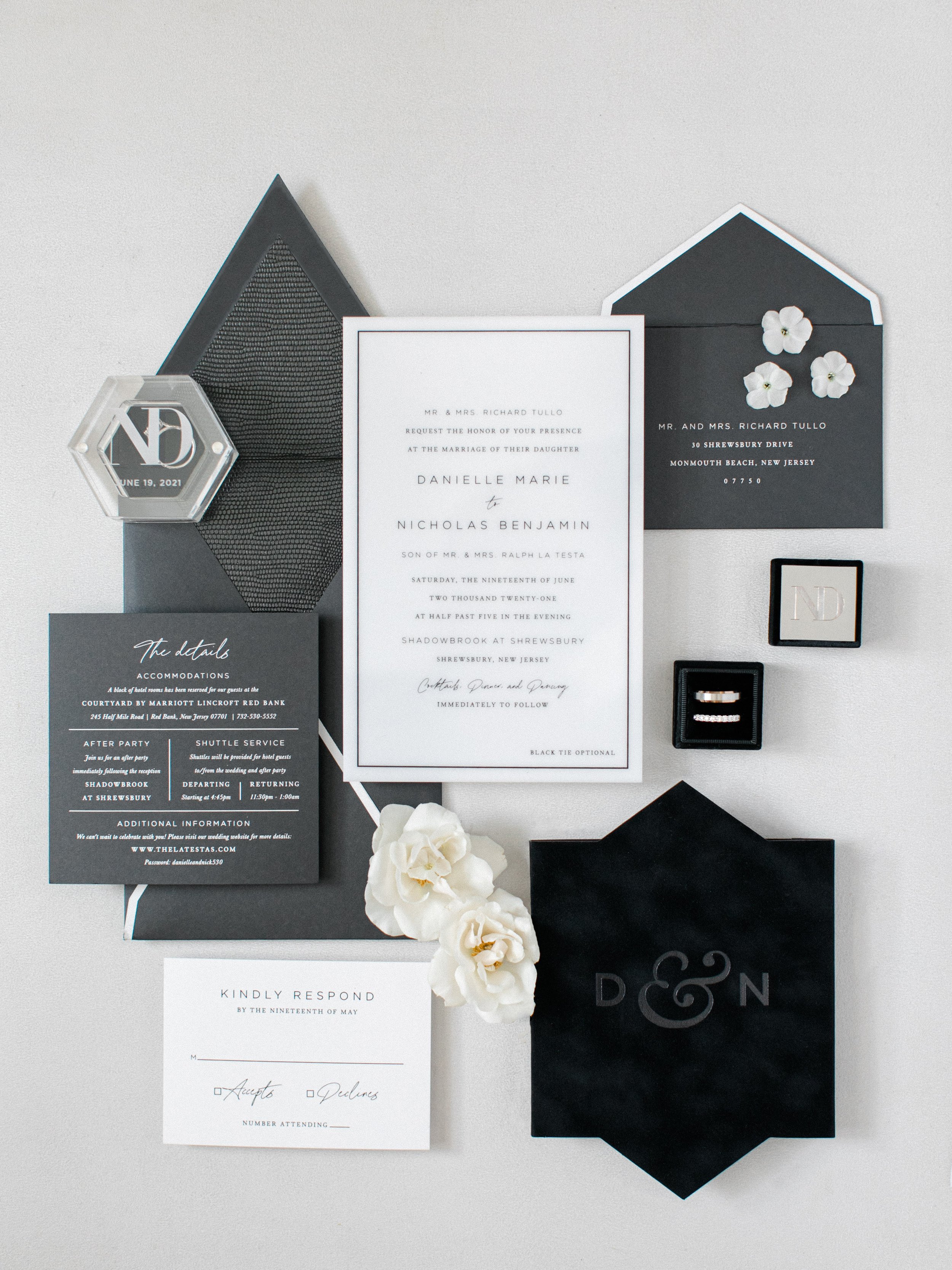 Black and white wedding invitations, a black box with rings, white flowers, and reception details all arranged on a light gray background.