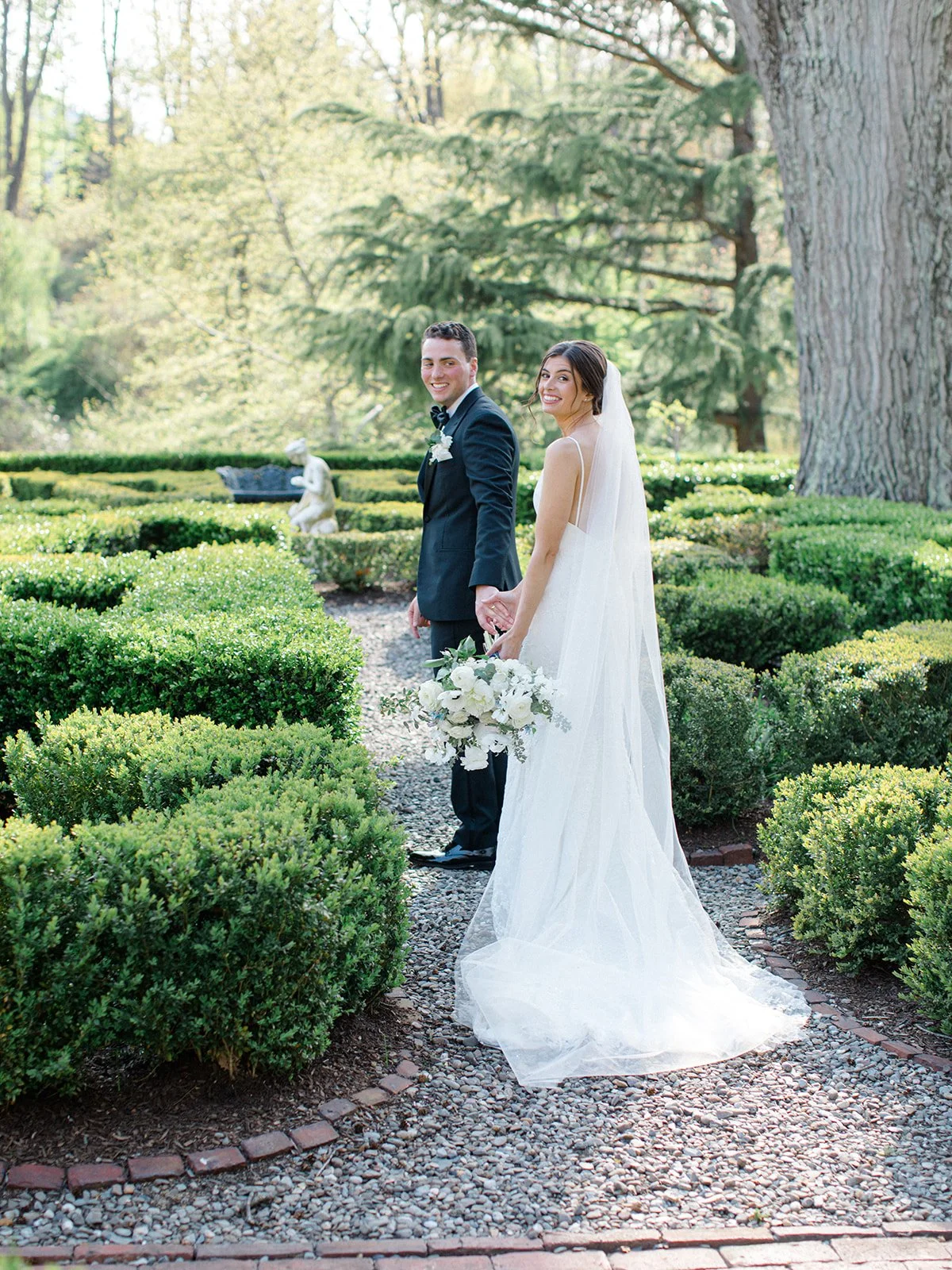 Bride and groom holding hands in a garden with manicured bushes, statues, and large trees in the background.