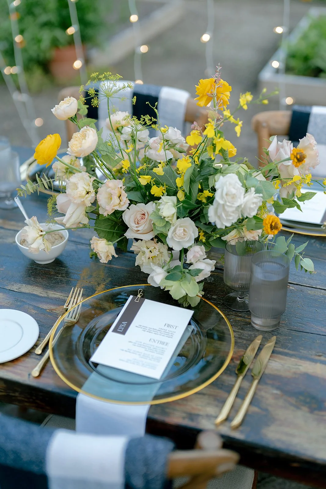 Elegant outdoor dining table decorated with a large floral centerpiece of white and yellow flowers, with gold cutlery, gray glasses, a menu, and a white table runner.