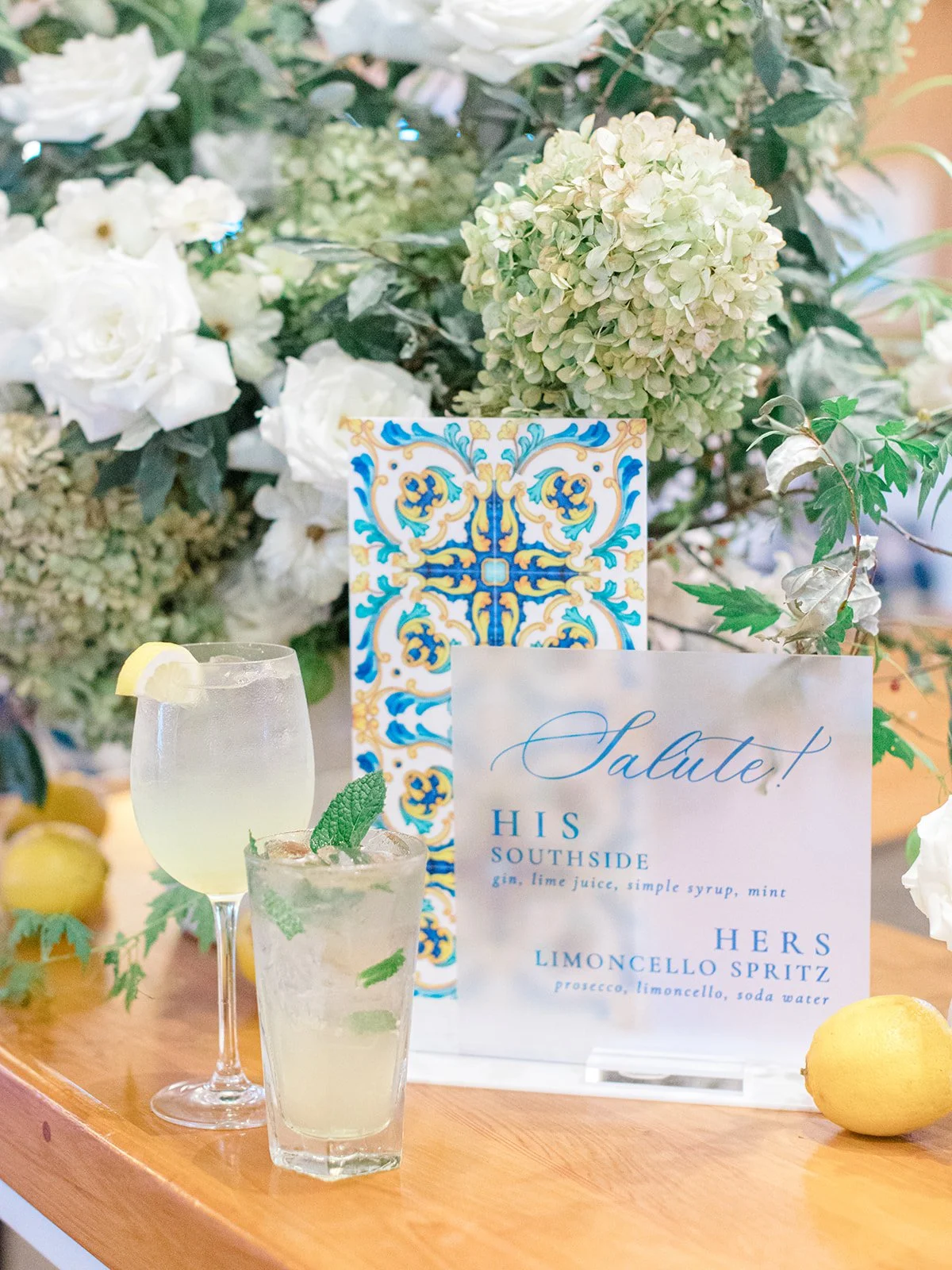 Two cocktails with lemon and mint garnishes placed on a wooden table, surrounded by white flowers and greenery, with a colorful tile on the background and a sign listing drink options.