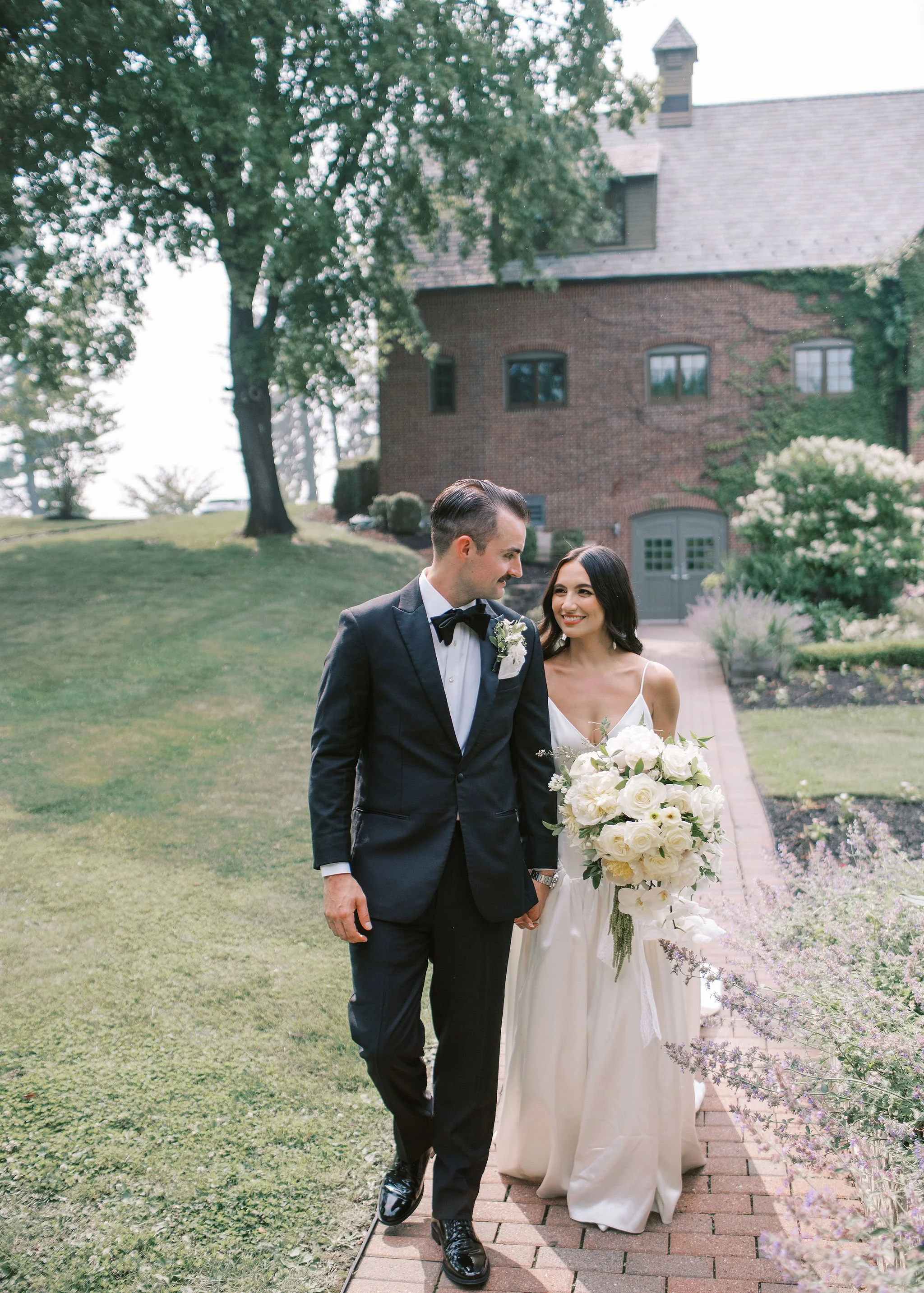 A bride and groom walk hand in hand on a brick path outdoors, with the bride holding a large white floral bouquet, smiling at each other. The groom is dressed in a black tuxedo with a bow tie, and the bride is wearing a white gown. There is greenery 