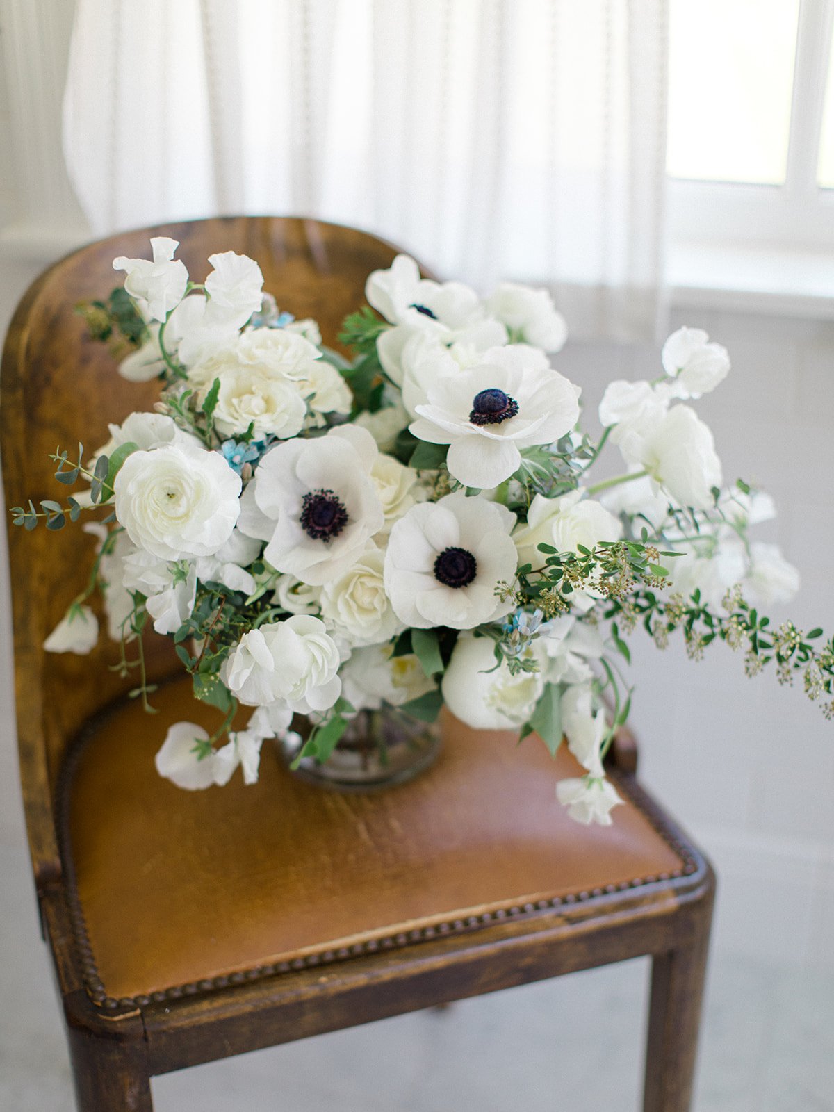 A white floral arrangement with anemones and greenery on a wooden chair near a window with sheer curtains.