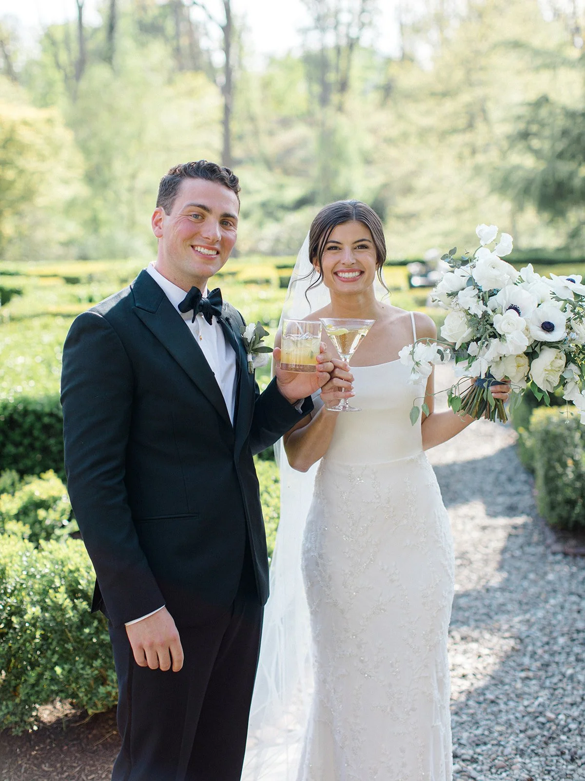 A newlywed couple dressed in wedding attire outdoors, holding drinks and smiling at the camera. The groom is wearing a black tuxedo with a bow tie, and the bride is wearing a white wedding dress and holding a bouquet of white flowers.