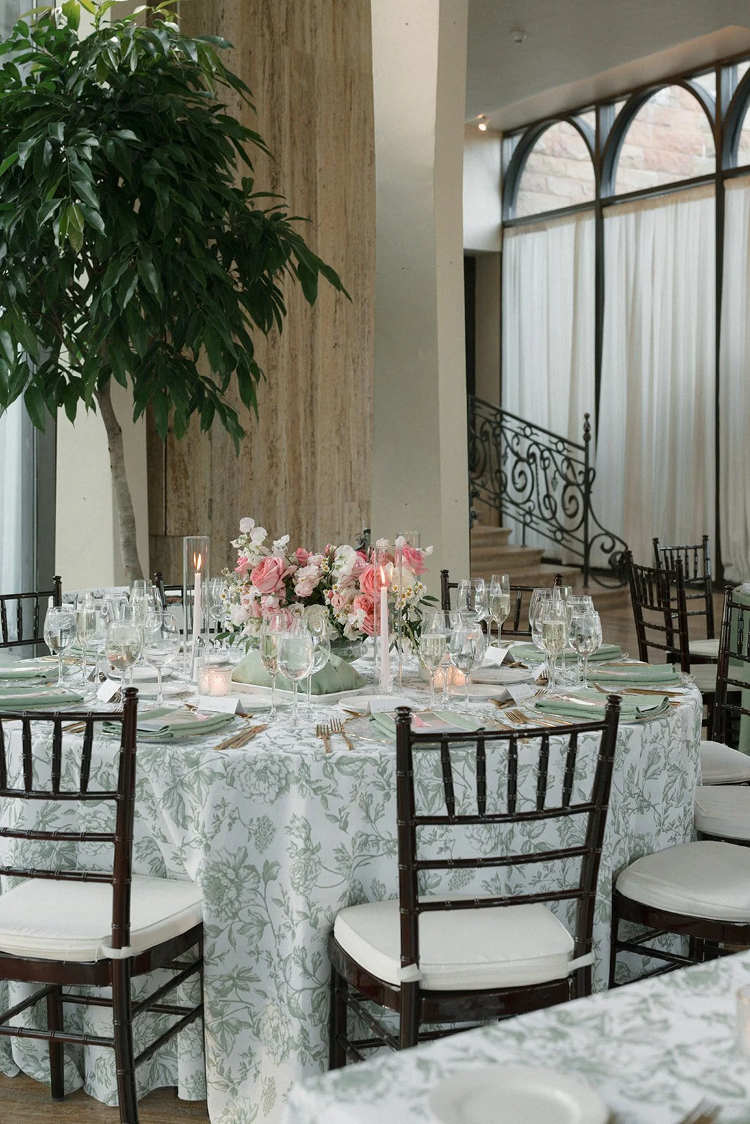 Elegant banquet table set with floral centerpiece, candles, glassware, and tailored place settings in a bright room with large windows, curtains, and decorative iron staircase.