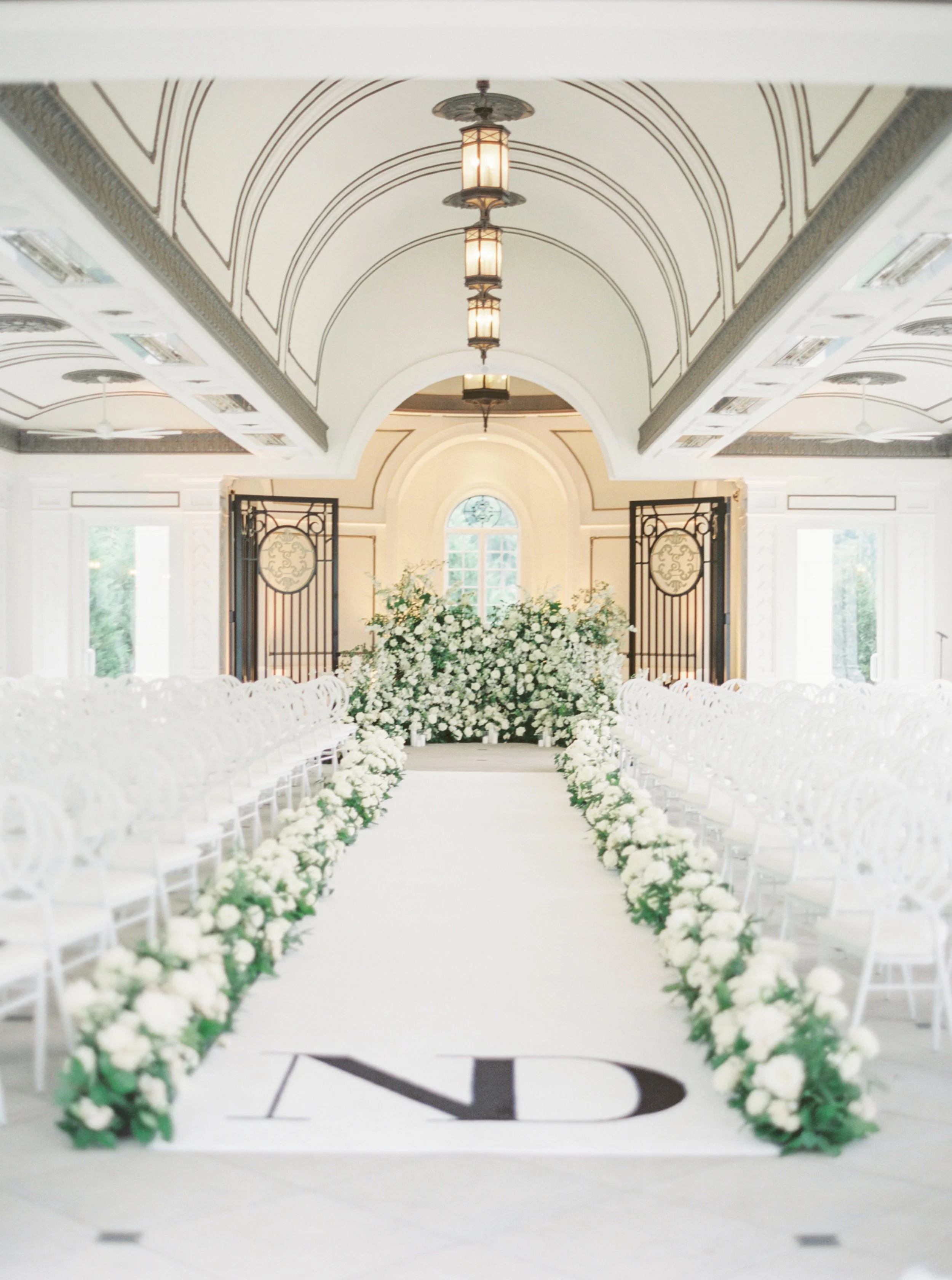 Elegant indoor wedding ceremony setup with a white aisle flanked by white flowers, white chairs, ornate decor, and a floral backdrop in a bright, airy room.