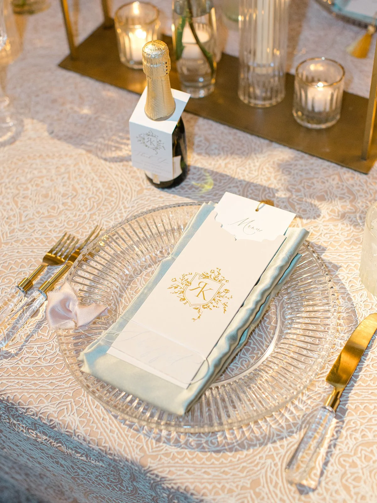 Elegant table setting with a clear glass charger plate, a folded napkin, a menu card with gold emblem, gold cutlery, a champagne bottle with a decorative sleeve, and candles in glass holders on a lace tablecloth.