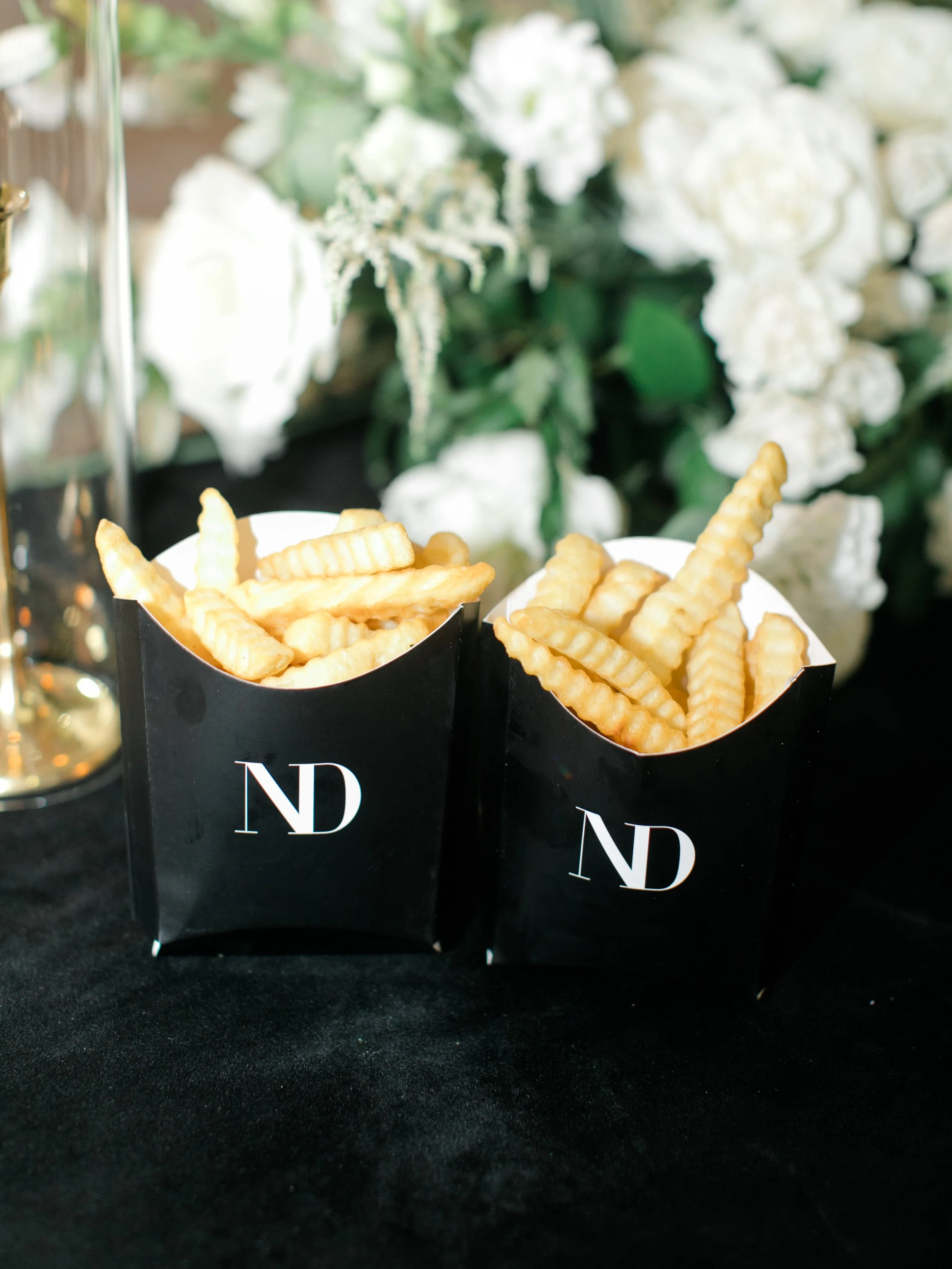 French fries in black boxes with white lettering, set on a black surface with white flowers and green foliage in the background.