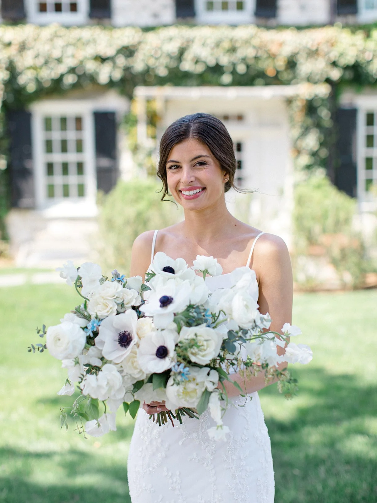 A smiling woman in a white dress holding a large bouquet of white and purple flowers outdoors.