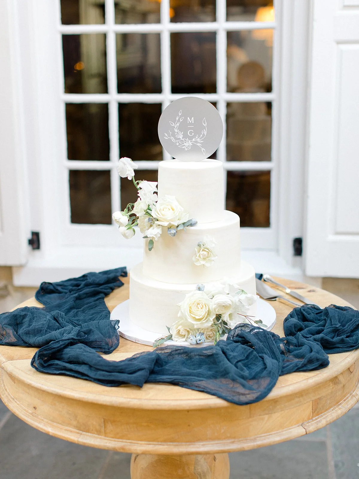 A three-tier white wedding cake decorated with white roses and greenery, topped with a round monogrammed cake topper with the initials M and G, on a wooden table with blue fabric and cake serving utensils, in front of a large window.