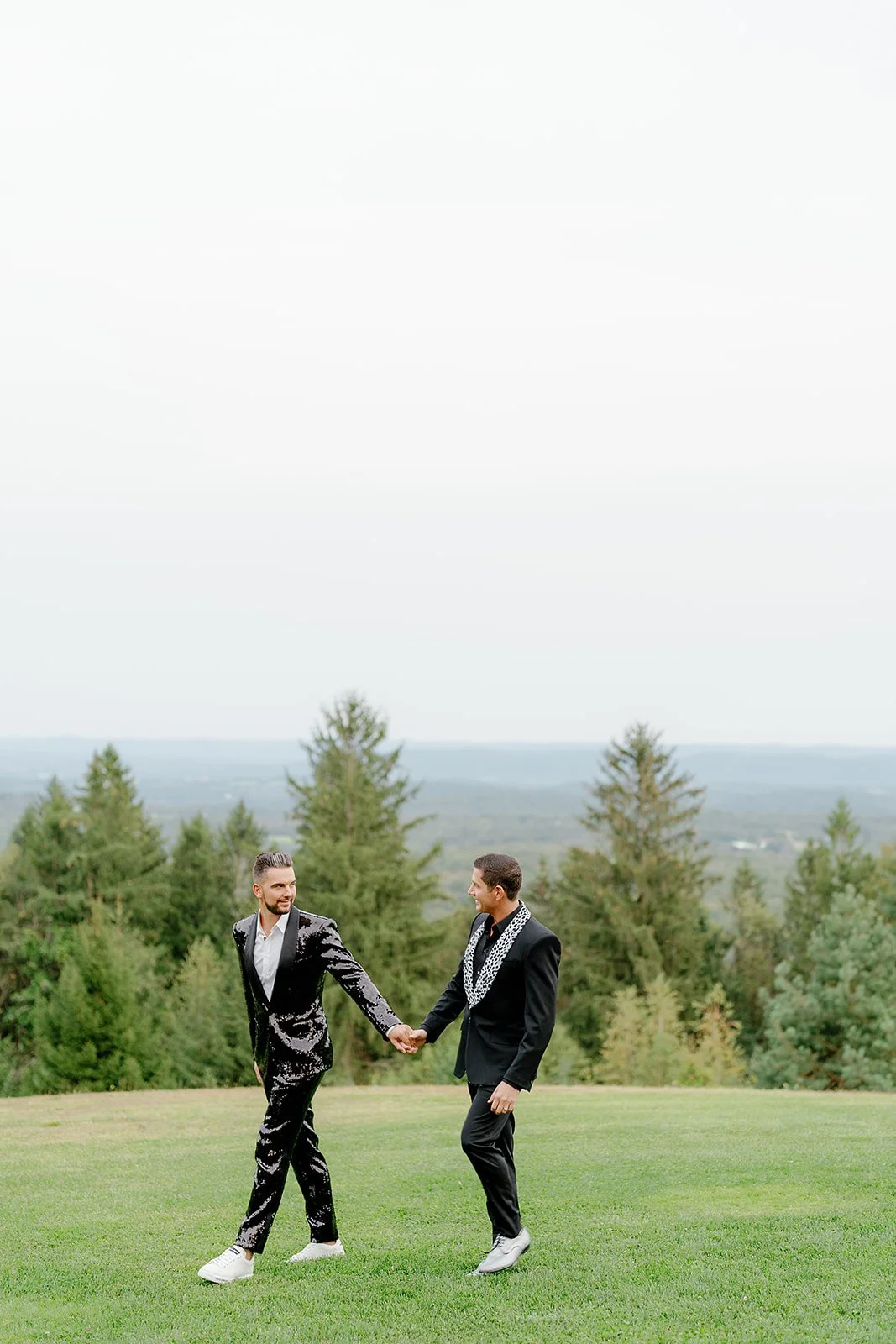 Two men in suits holding hands, walking on a grassy area with trees and a cloudy sky in the background.