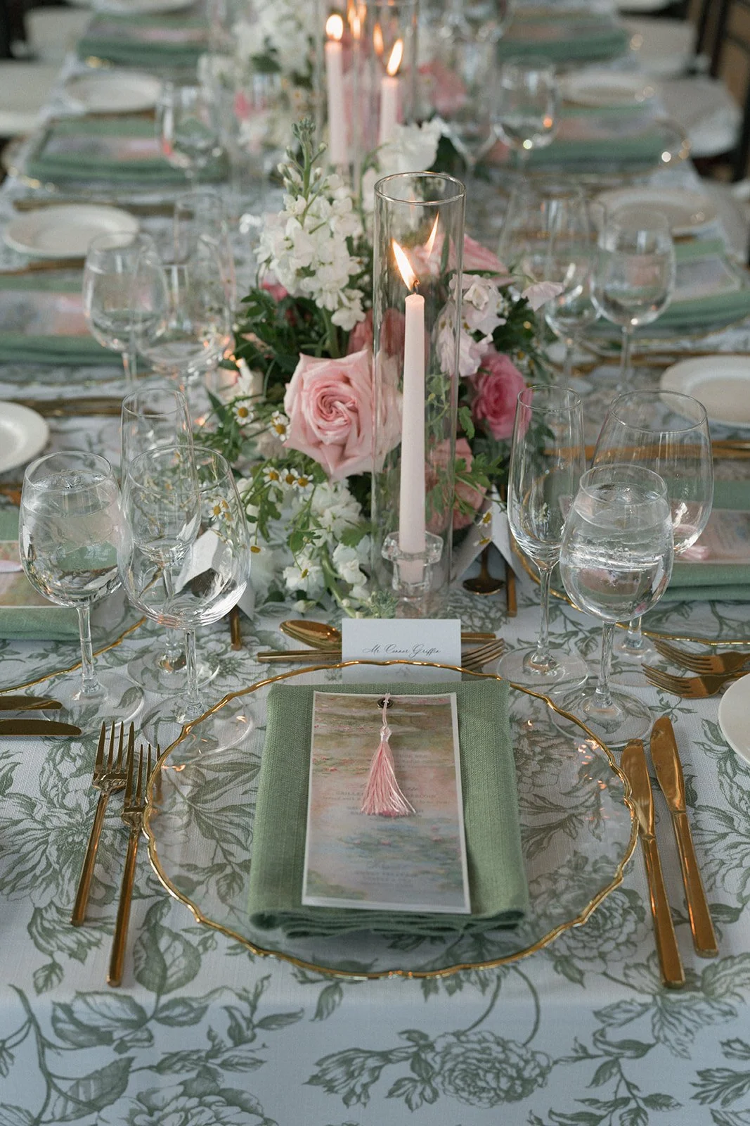Elegant dining table with floral centerpiece, pink roses, white flowers, and a tall glass candlestick with a white candle. Gold flatware, clear glassware, green napkins, and a patterned tablecloth with green leaves design. A menu with a pink tassel i