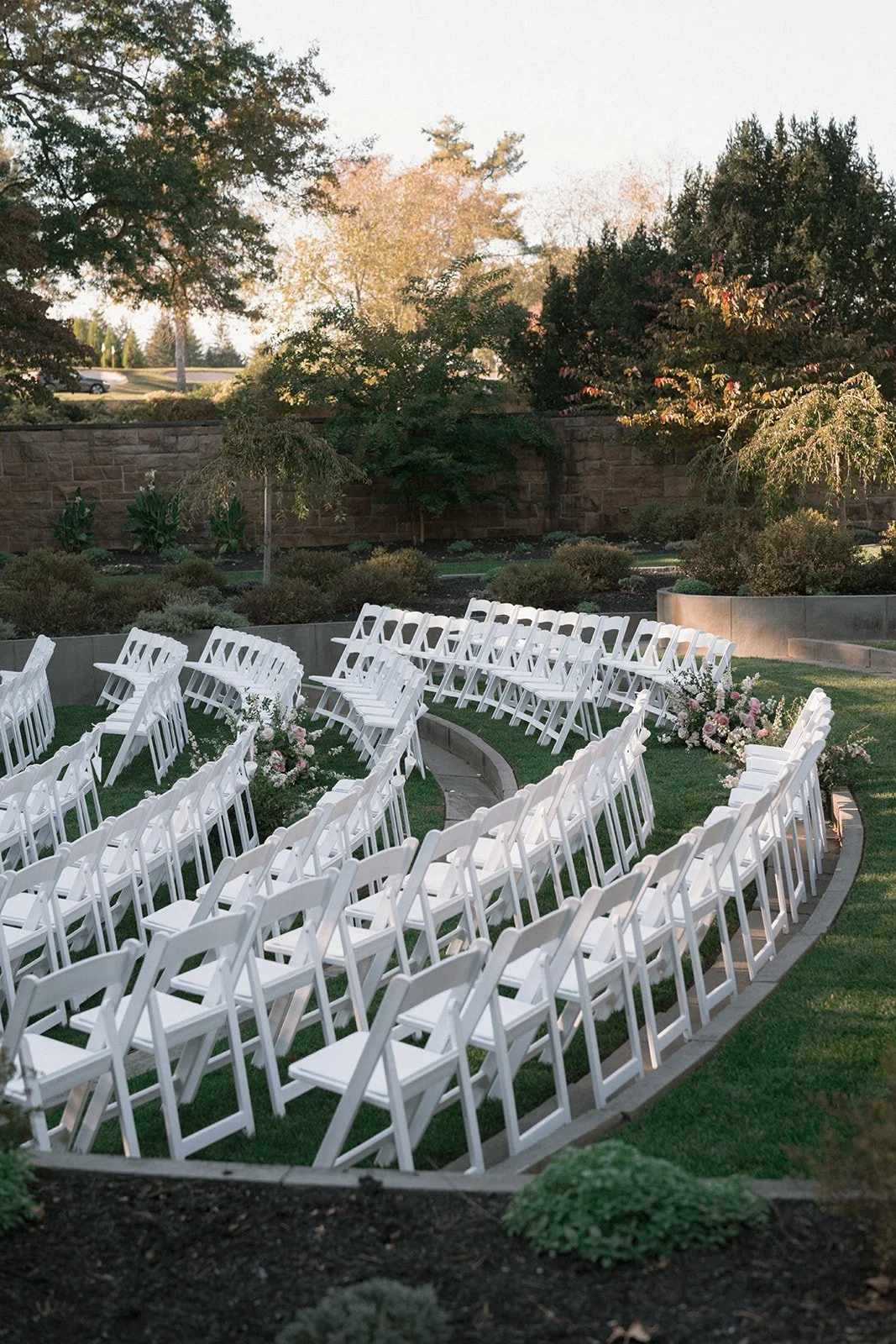 White chairs arranged in a semi-circle for an outdoor wedding ceremony, with floral decorations along the ground and greenery in the background.