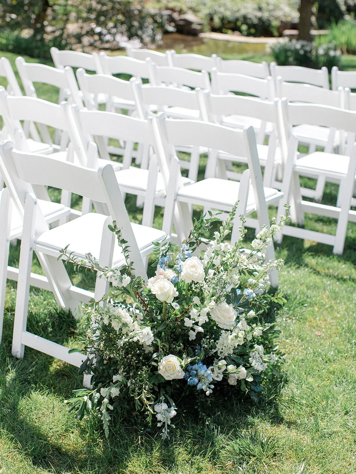 White folding chairs arranged outdoors with a floral arrangement of white and light blue flowers at the front.