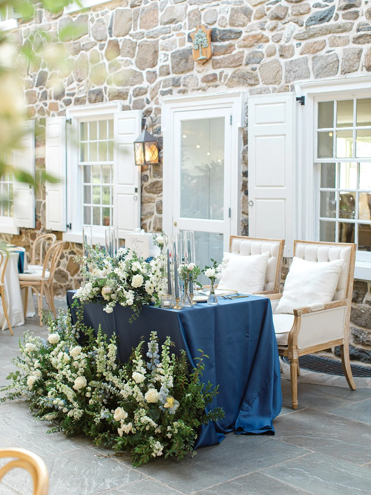 Decorated table with white floral arrangements, candles, and navy tablecloth in front of stone house with white shutters, two chairs with white cushions, and a wall-mounted lantern light fixture.