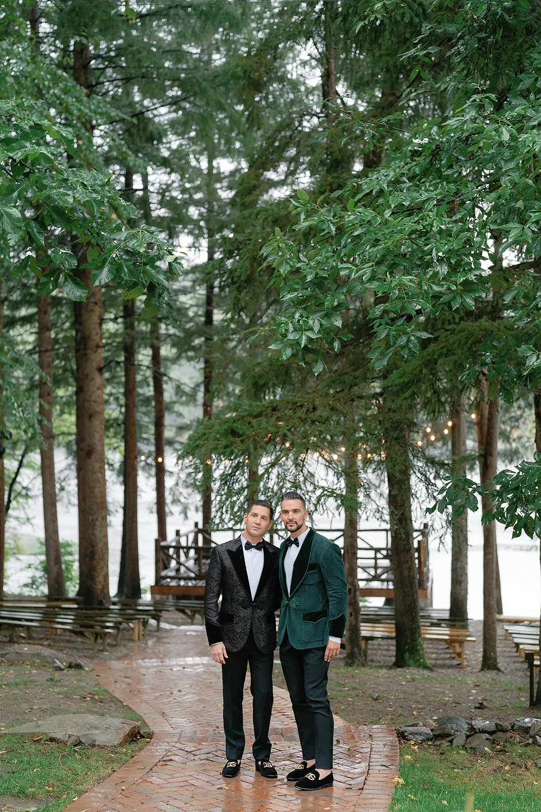 Two men standing together on a brick path surrounded by greenery in a forested area, dressed in tuxedos with bow ties, under string lights.