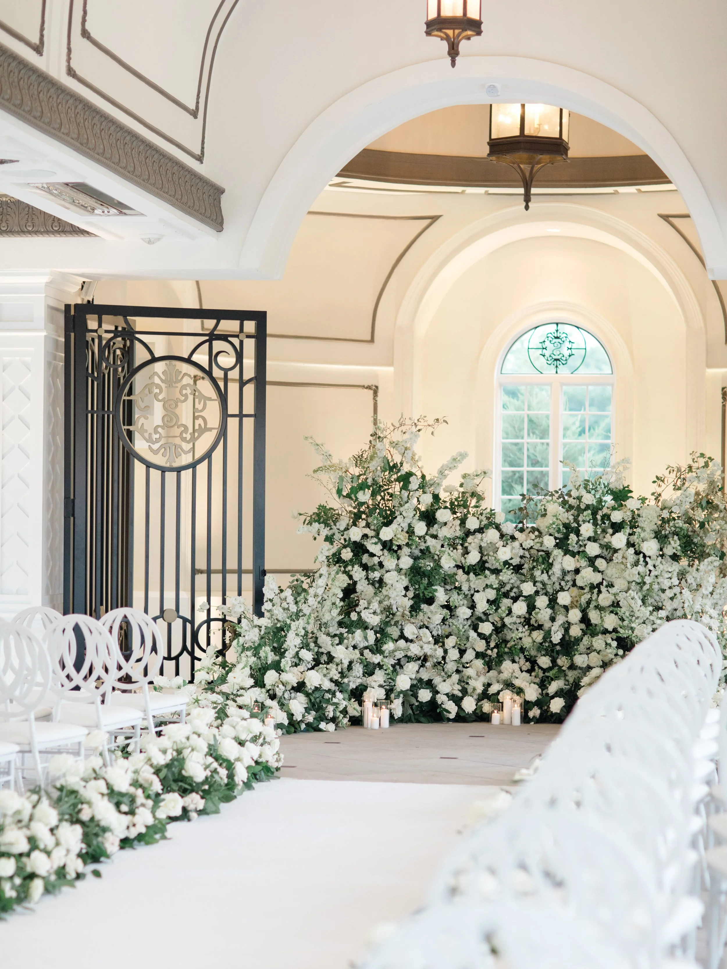 Elegant wedding aisle decorated with white flowers and candles, inside a bright, decorated room with arched windows and ornate lighting fixtures.