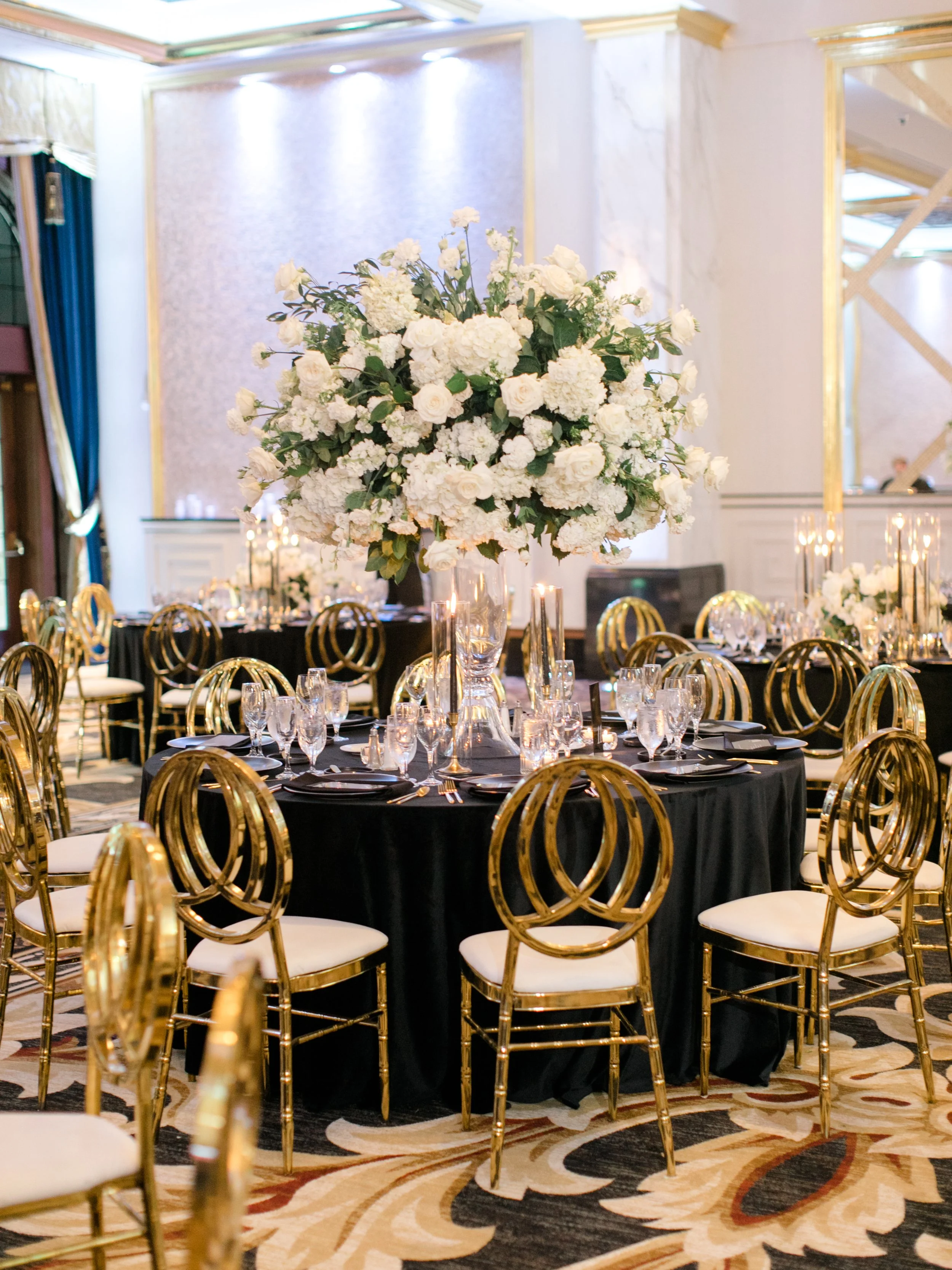 A banquet table decorated with a large white floral centerpiece, glasses, and black tablecloths, set in an elegant event hall with gold chairs and ornate wall details.