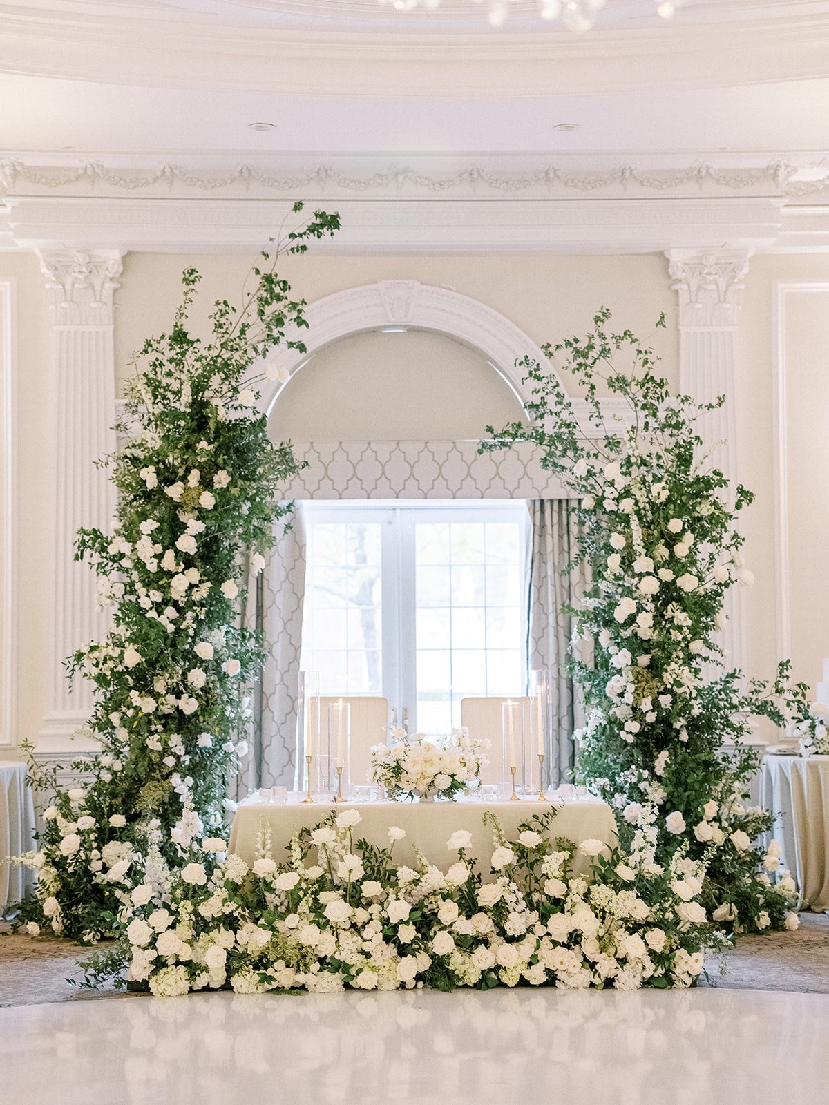 Elegant indoor wedding reception setup with a floral arch, white flowers, candles, and a cream-colored table near large window.