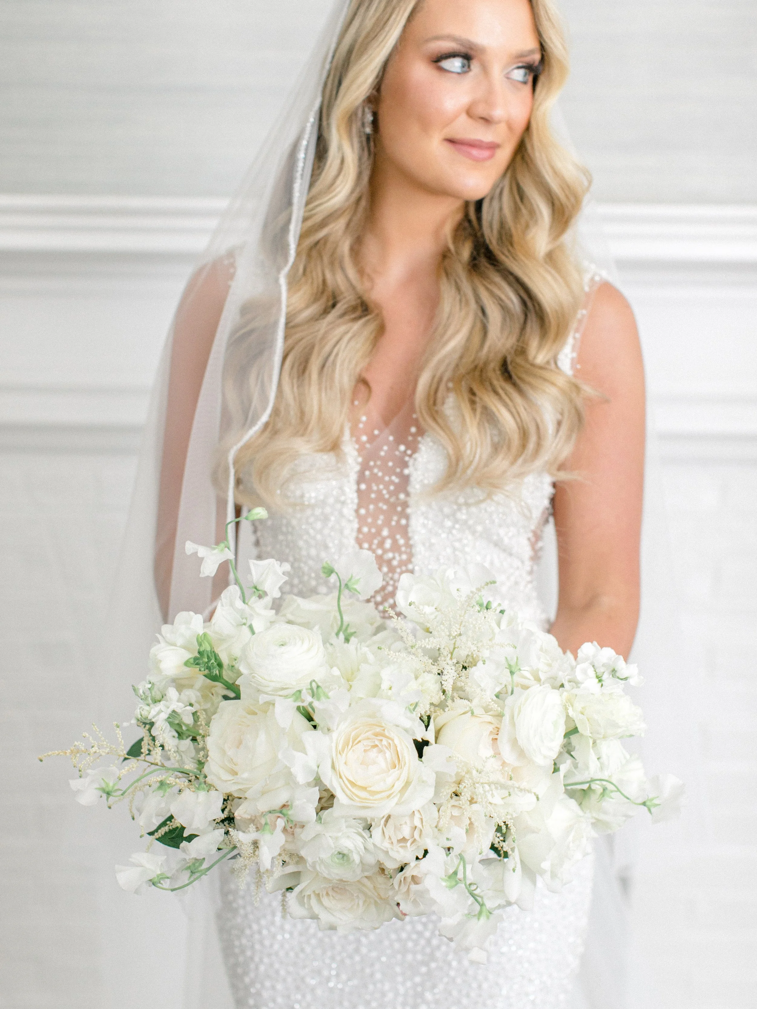 A bride with long blonde wavy hair, wearing a white wedding dress with pearl embellishments and a sheer, beaded neckline, holding a large bouquet of white flowers, standing in a bright room.