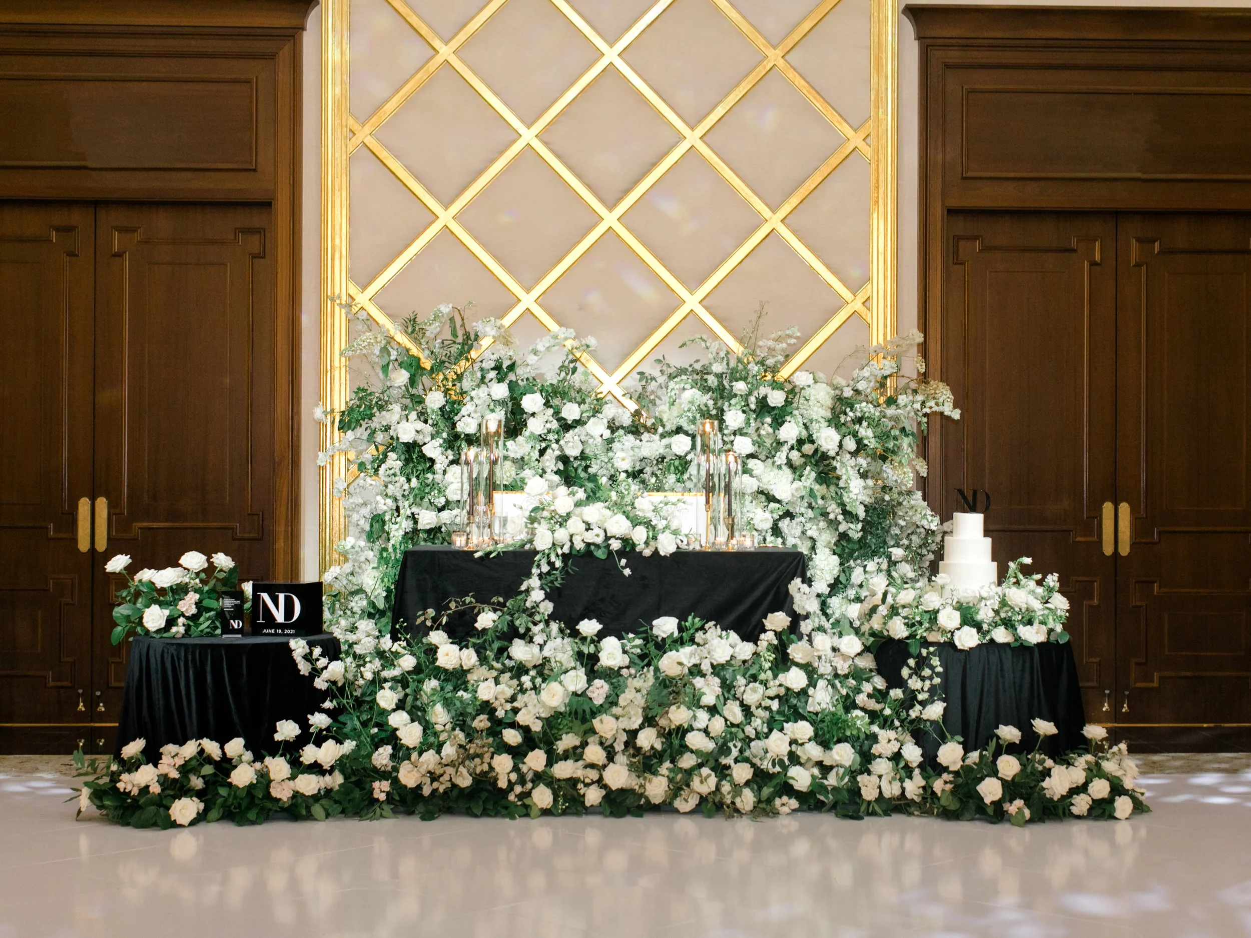Elegant wedding cake table decorated with white flowers and greenery, with a black tablecloth, set against a gold and pink patterned backdrop, wooden doors on either side.