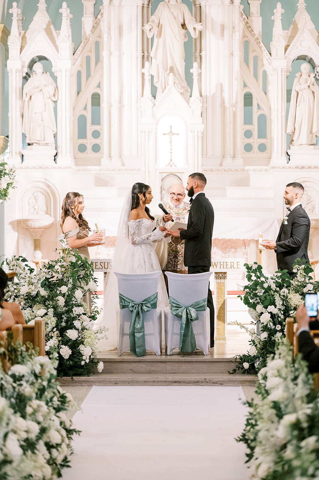 A wedding ceremony inside a church with a bride and groom exchanging vows, surrounded by flowers and an altar with religious statues and a crucifix in the background.