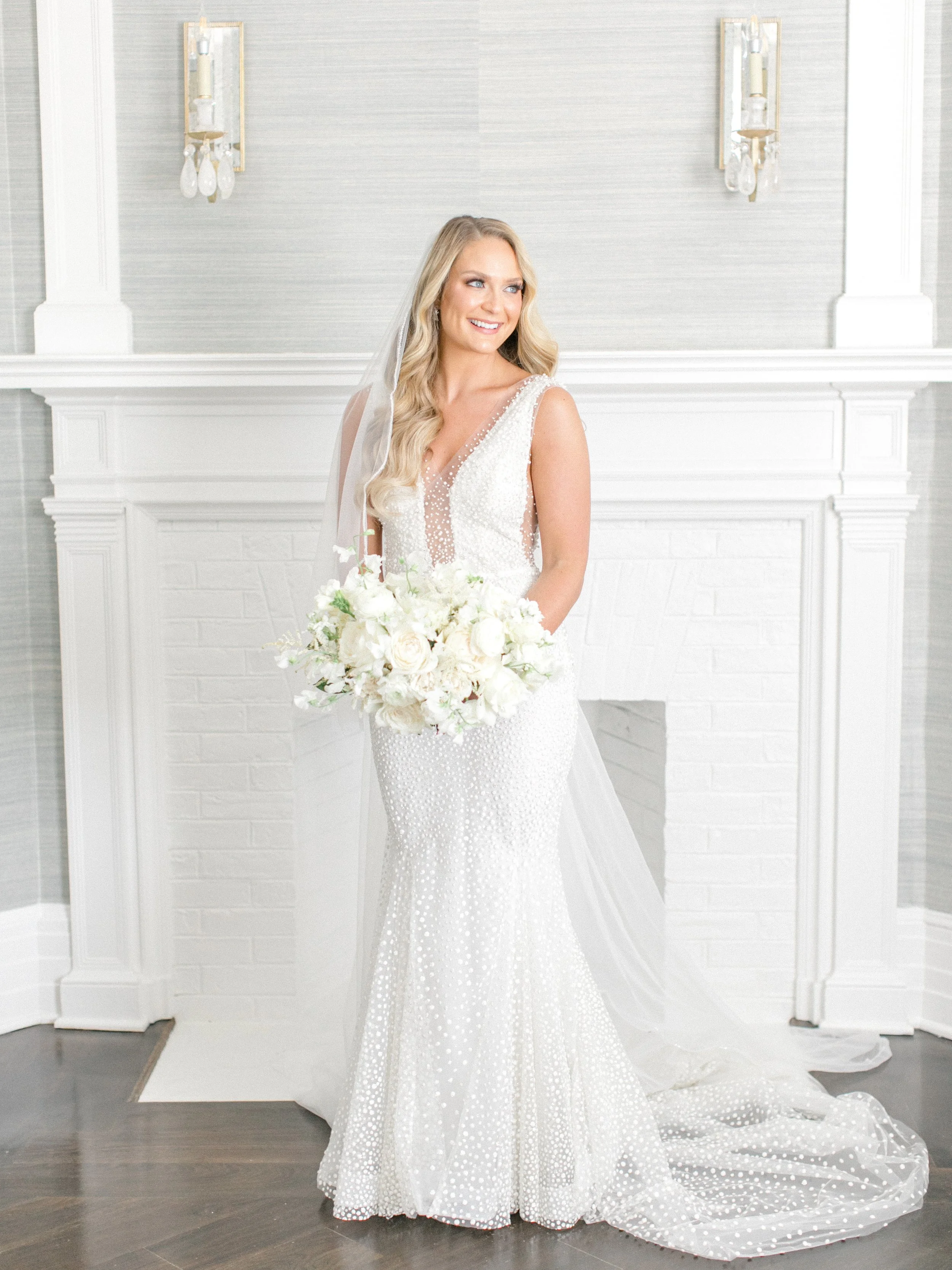A bride in a white wedding gown holding a bouquet of white flowers, standing in front of a white brick fireplace with two wall sconces.