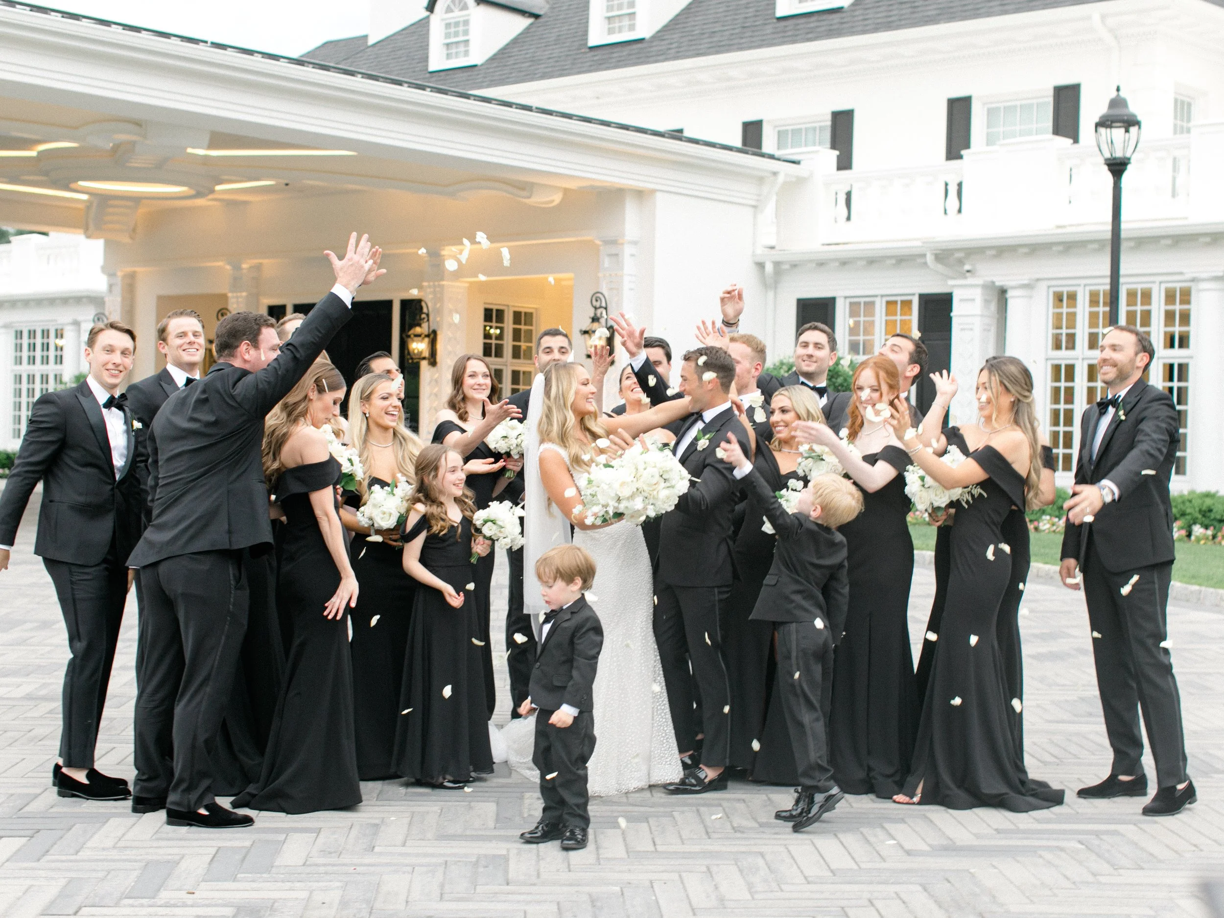 A bride and groom surrounded by wedding party, throwing confetti outside a white building with large windows and black shutters.