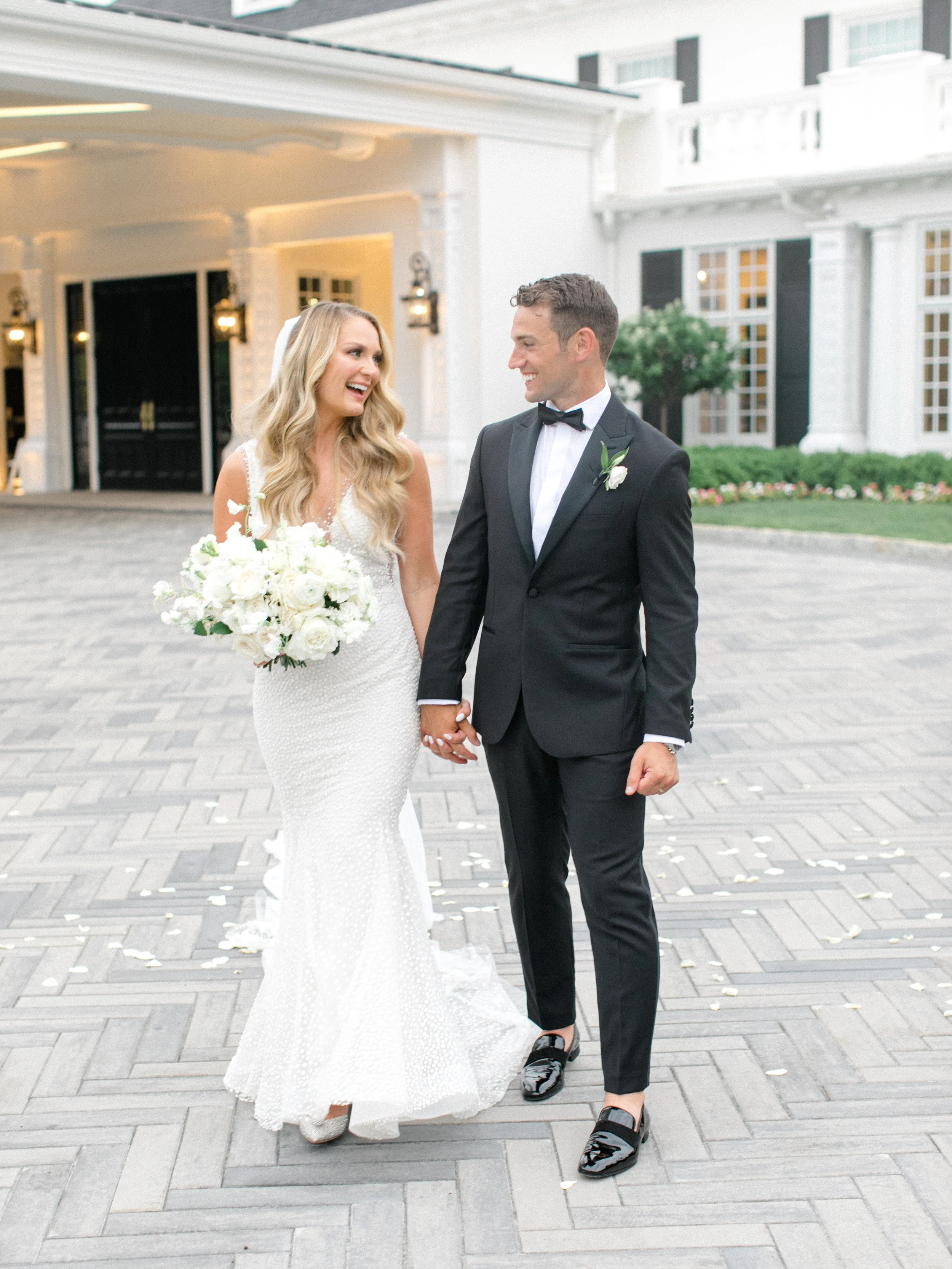 A bride and groom holding hands and smiling at each other outside a white building with large windows and a driveway, the bride in a white wedding dress holding a bouquet of white flowers, the groom in a black tuxedo with a bow tie.