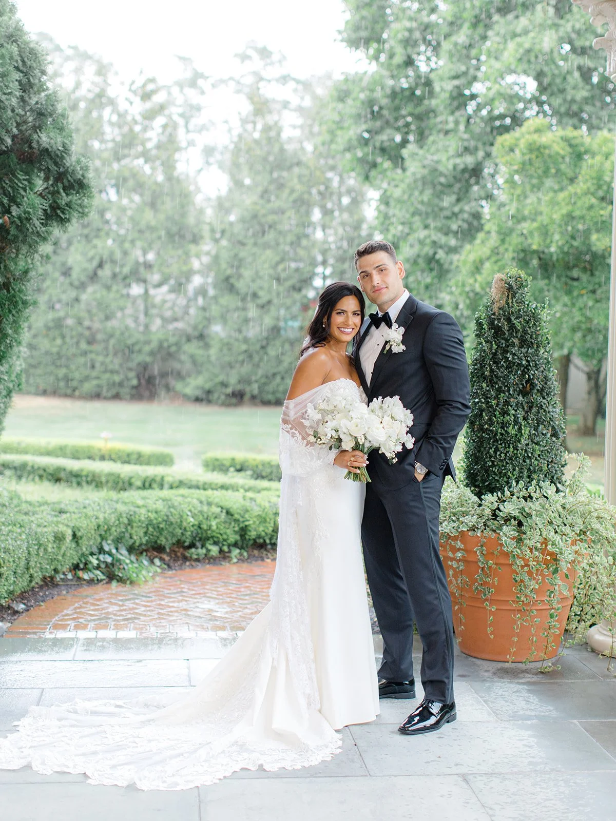 A newlywed couple in wedding attire, the bride in an off-shoulder white gown holding a bouquet of white flowers, and the groom in a black tuxedo with a bow tie, standing on a porch with greenery background and a large potted plant nearby.