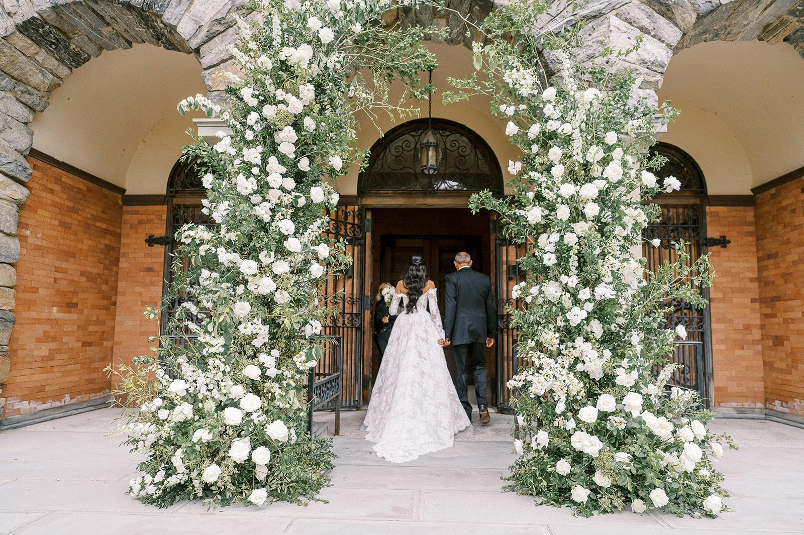 A bride and groom walking through an ornate floral archway at their wedding, with the bride in a long white dress and the groom in a dark suit, entering a building with brick walls and large wooden doors.