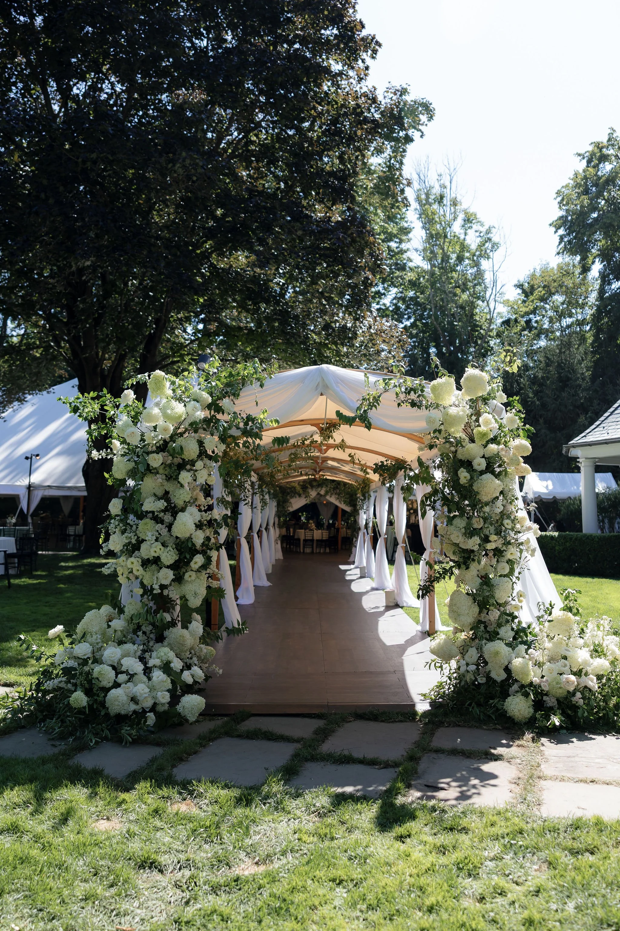 White floral archway with draped fabric set up outdoors for a wedding or event, surrounded by trees and a green lawn.