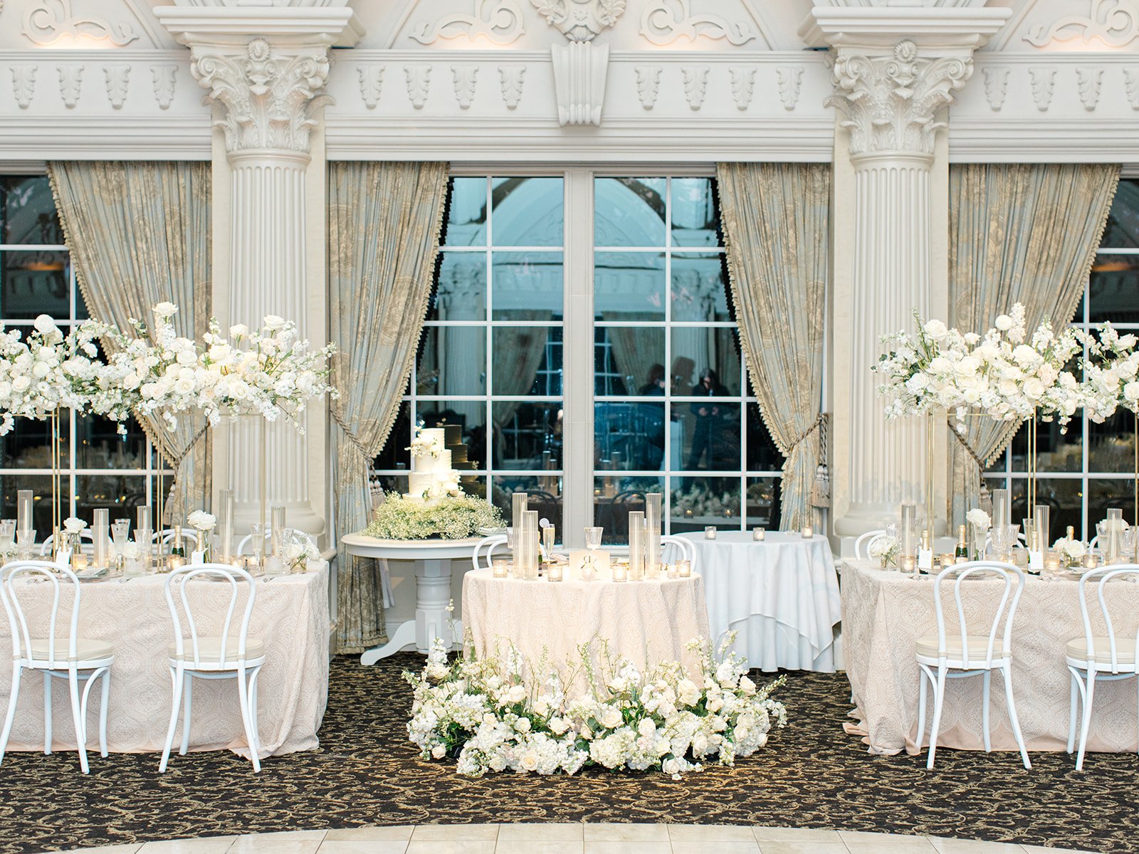 Elegant wedding reception setup featuring long tables with white tablecloths, floral centerpieces with white flowers, candles, and tall floral arrangements. Large windows with ornate curtains and decorative columns are in the background, with a cake 