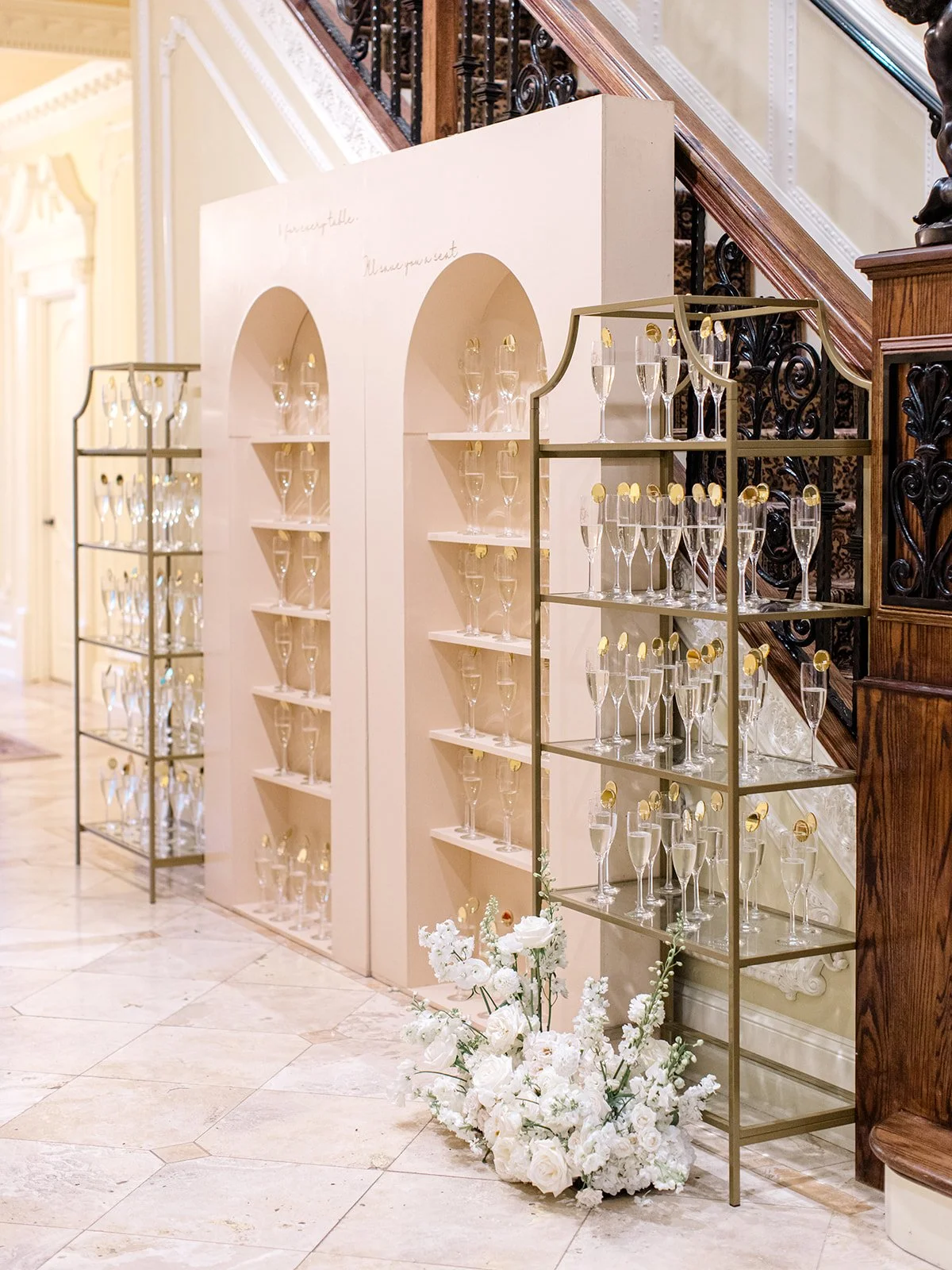 Display of champagne glasses with gold accents arranged on gold-colored shelving units in an elegant interior setting with marble flooring and floral arrangements.