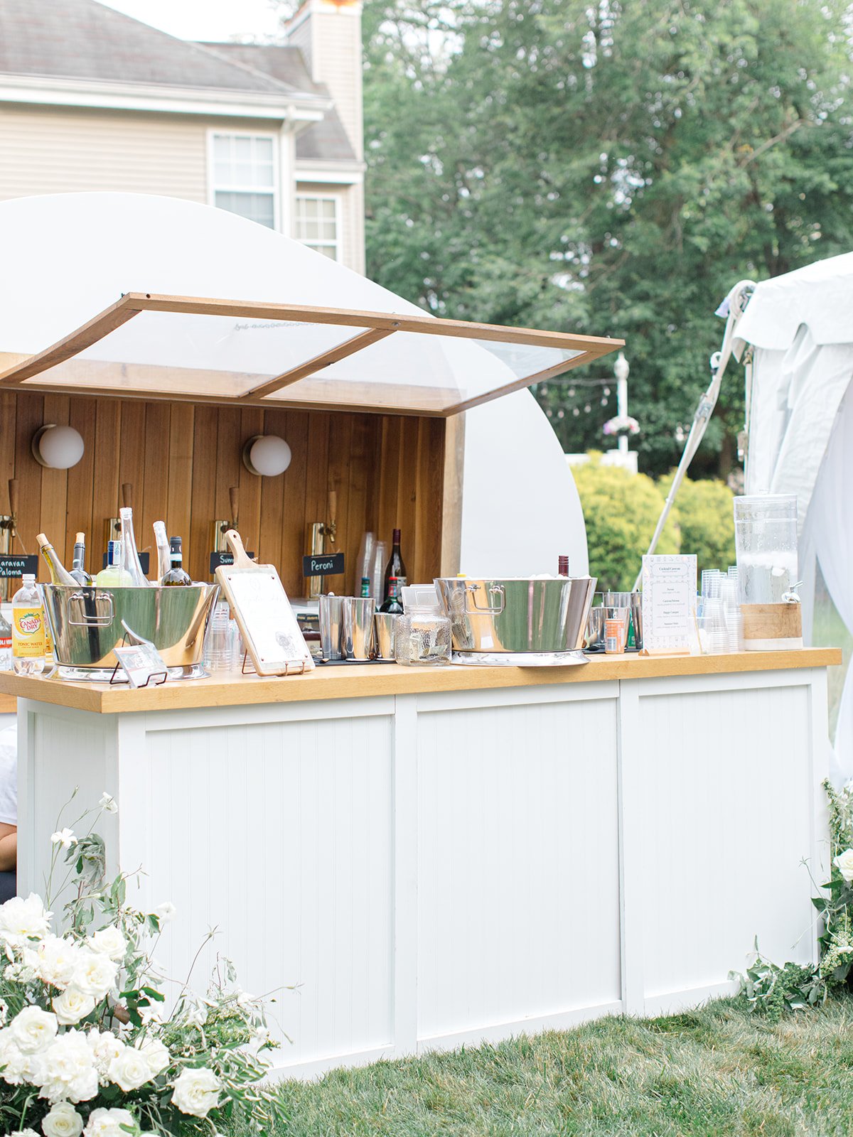 Outdoor bar setup with liquor bottles, ice buckets, and cups in a garden setting with trees, a white tent, and a house in the background.