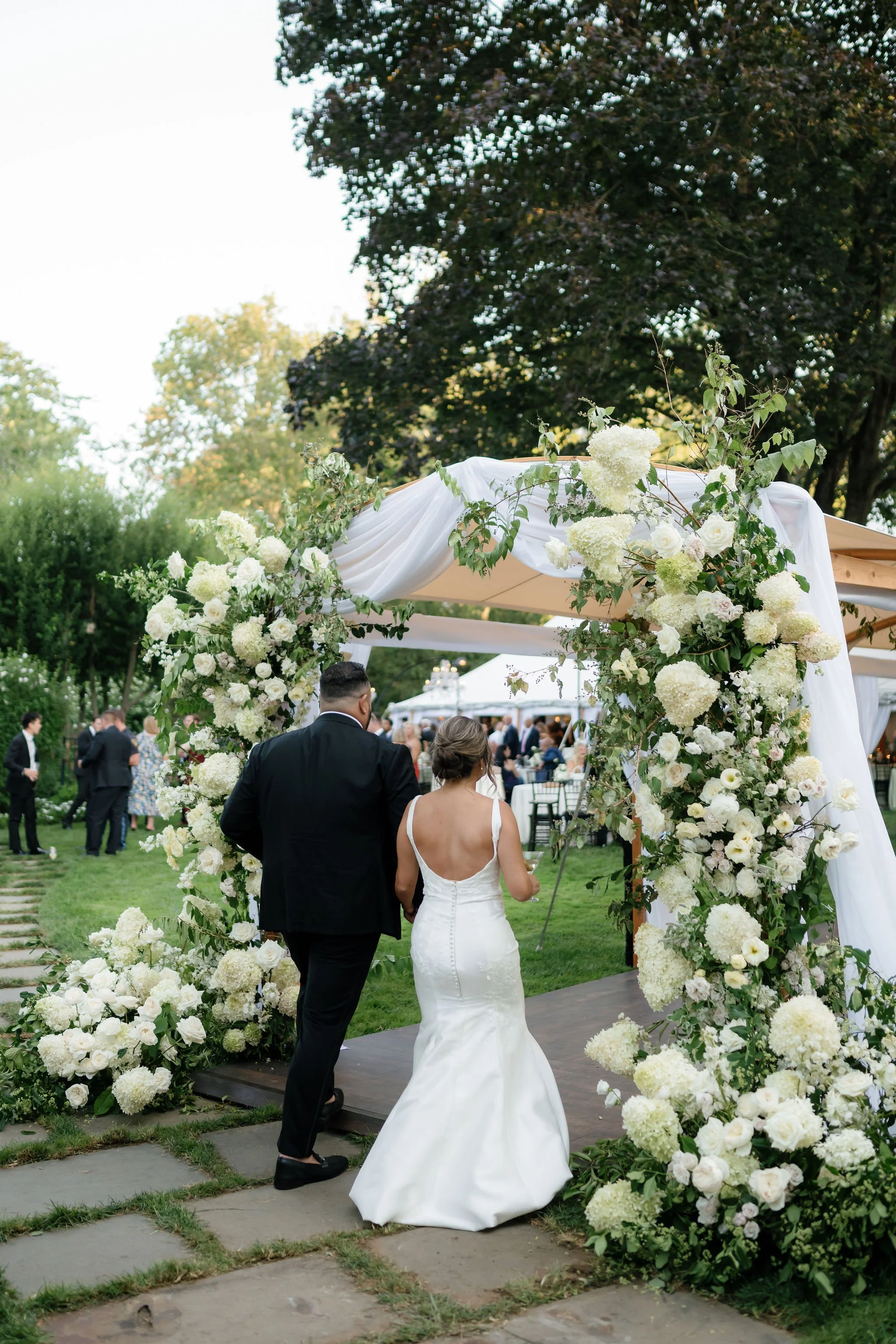 A bride in a white wedding dress and a groom in a black suit walking through a floral wedding arch on an outdoor garden wedding venue.