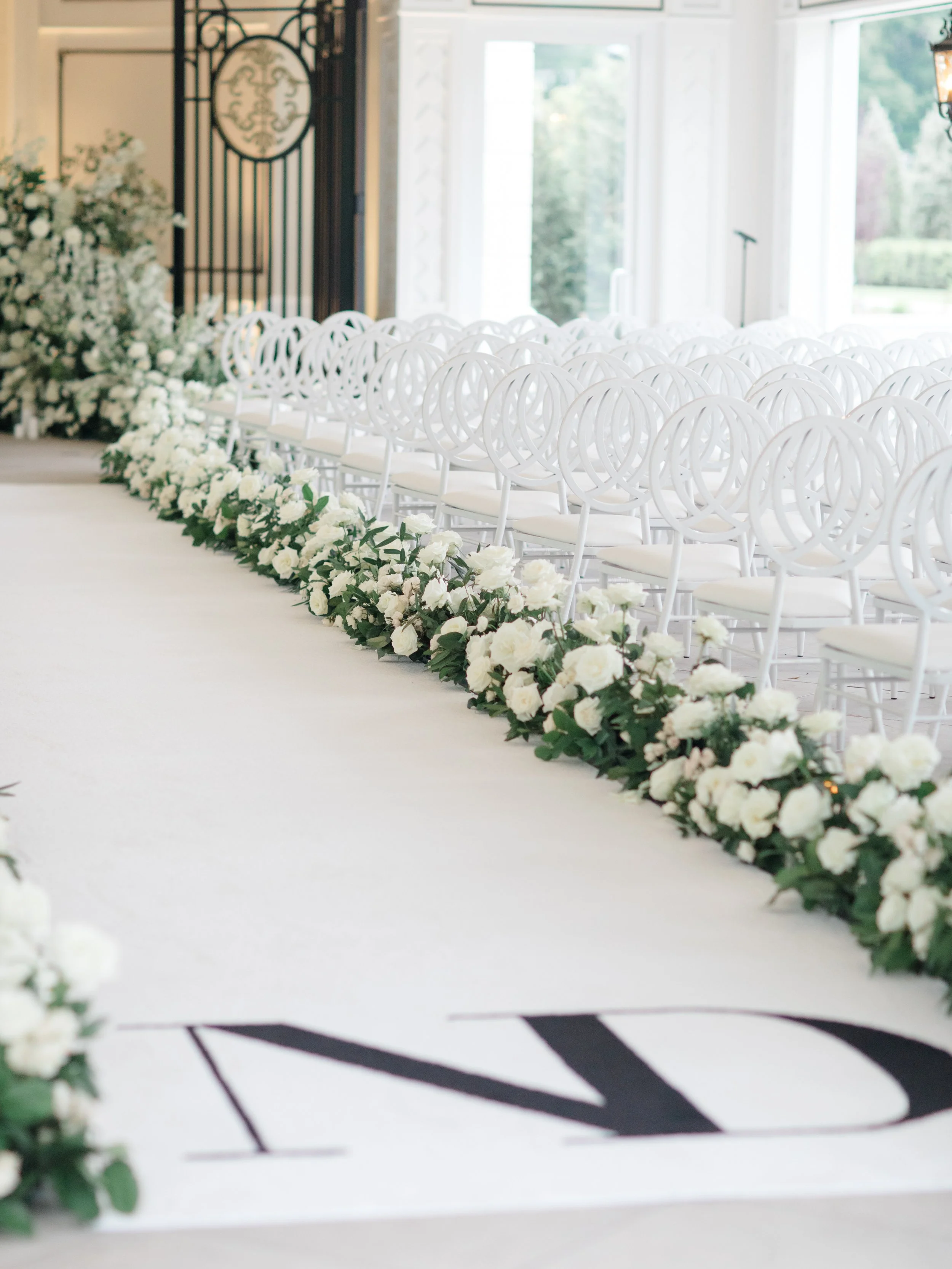 Wedding ceremony setup with white chairs arranged in rows and decorated with white flowers along the aisle, inside a bright, elegant venue.