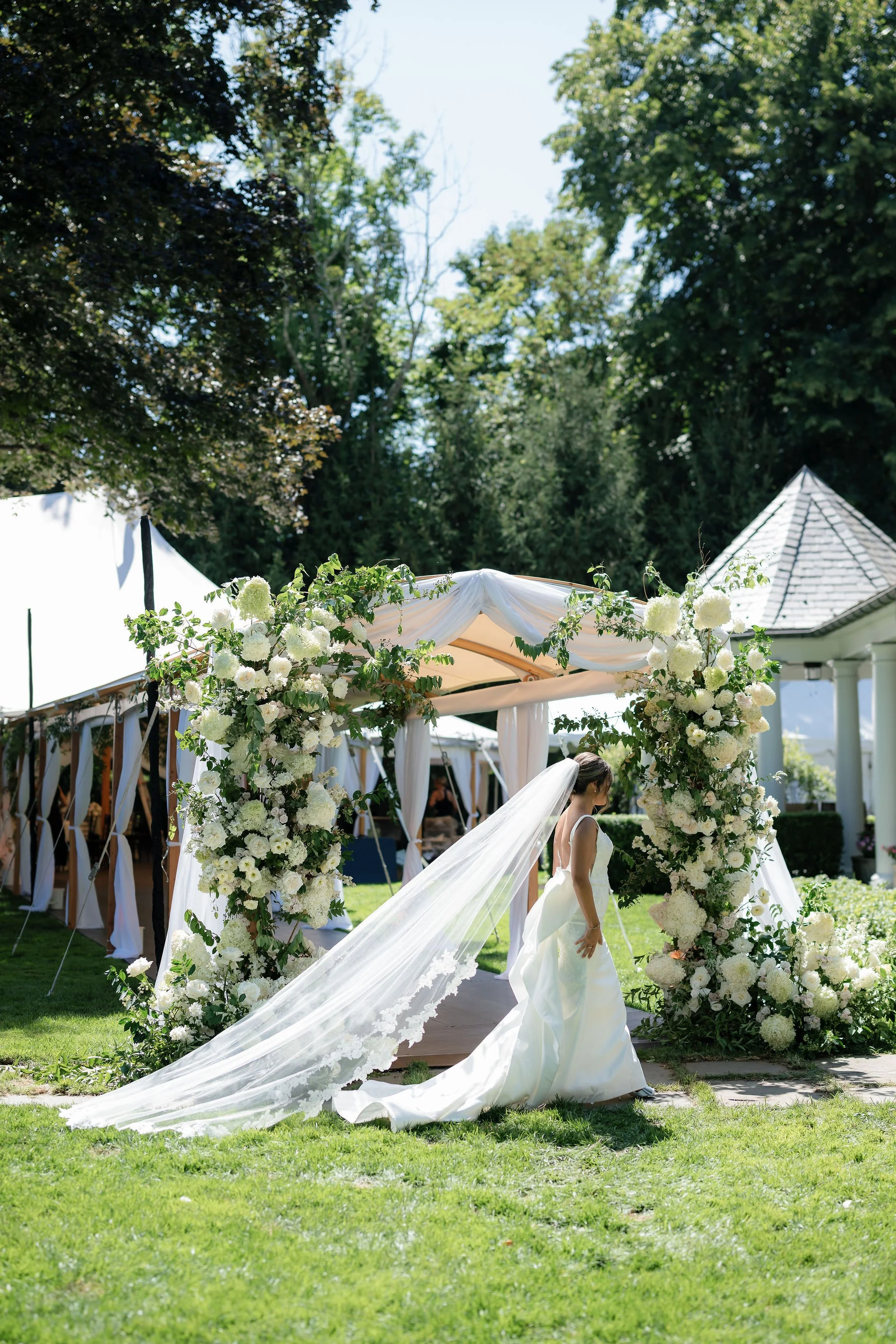A bride in a white wedding dress and long veil walking past a decorated archway with white flowers at an outdoor wedding venue.