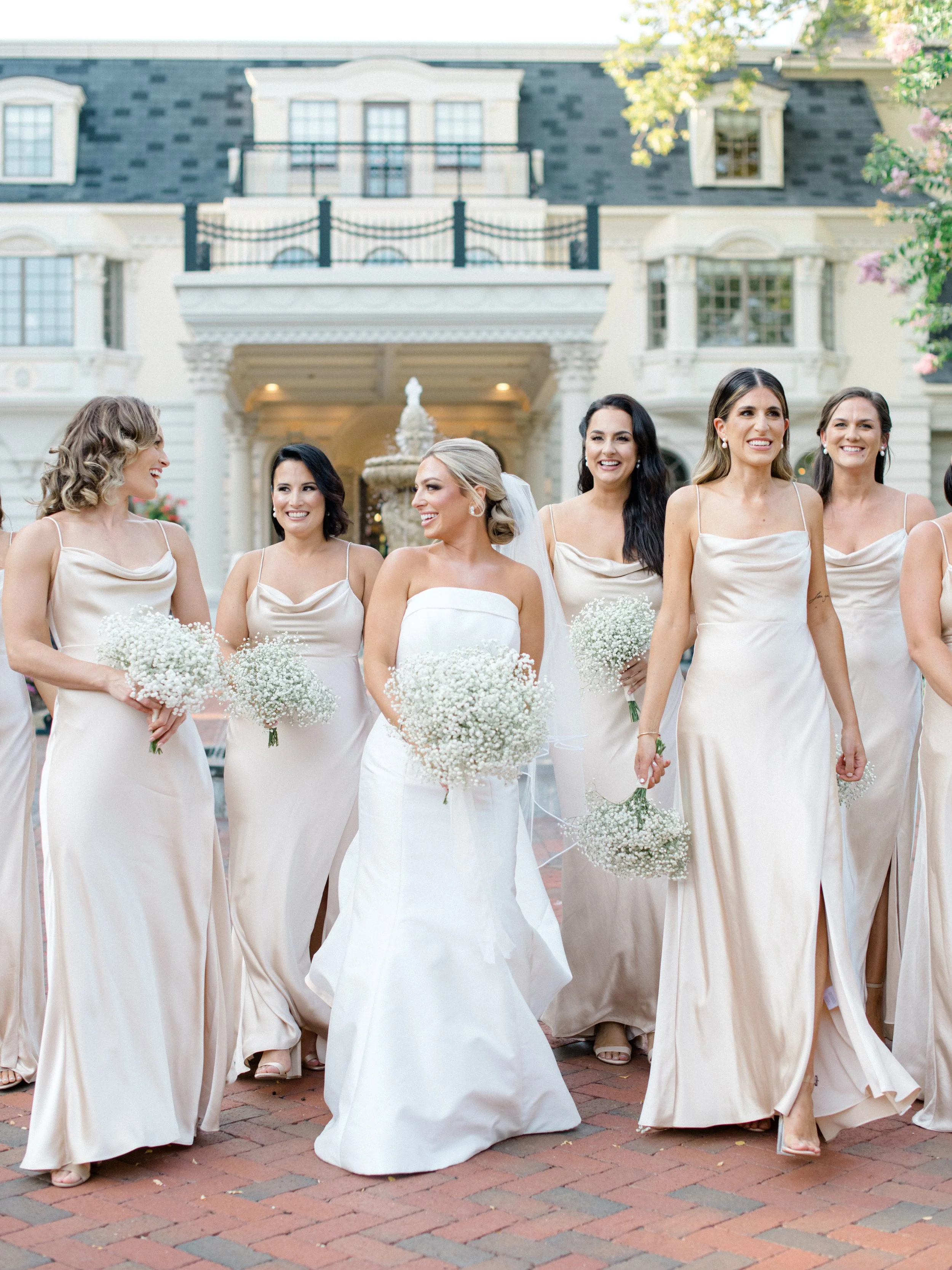 A group of seven women, including a bride in a white wedding dress, walking outside on a brick pathway in front of a large white mansion with a water fountain, surrounded by trees and pink flowers. The women are wearing matching light-colored dresses