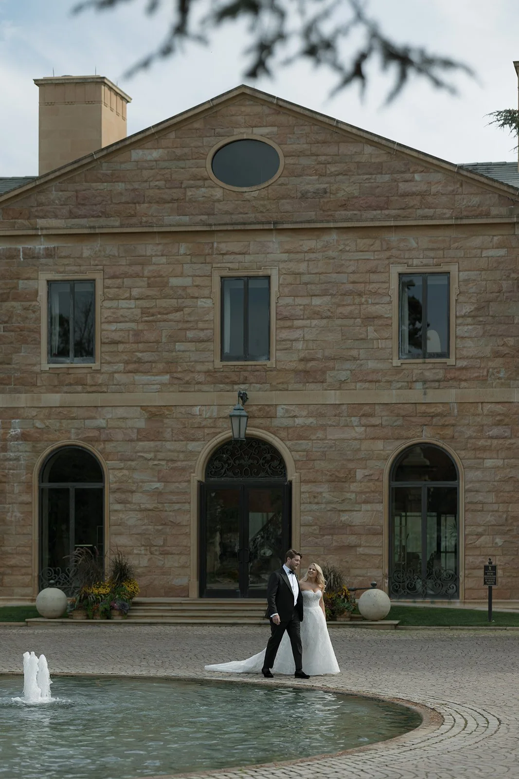 A bride and groom standing by a fountain in front of a large stone building, likely during their wedding.