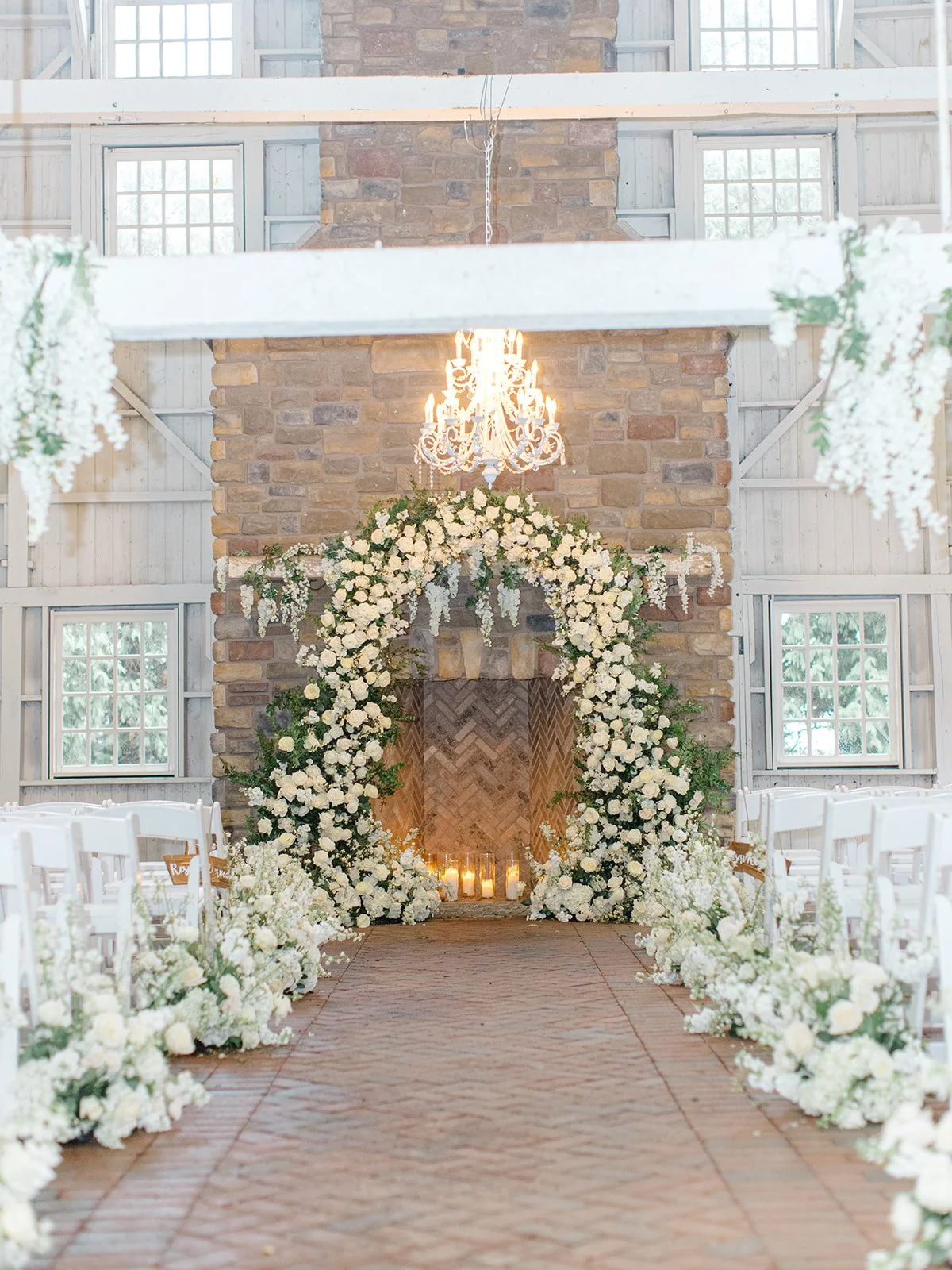 Wedding ceremony setup with white flower decorations, a floral arch, a brick fireplace, candles, a chandelier, and white chairs.