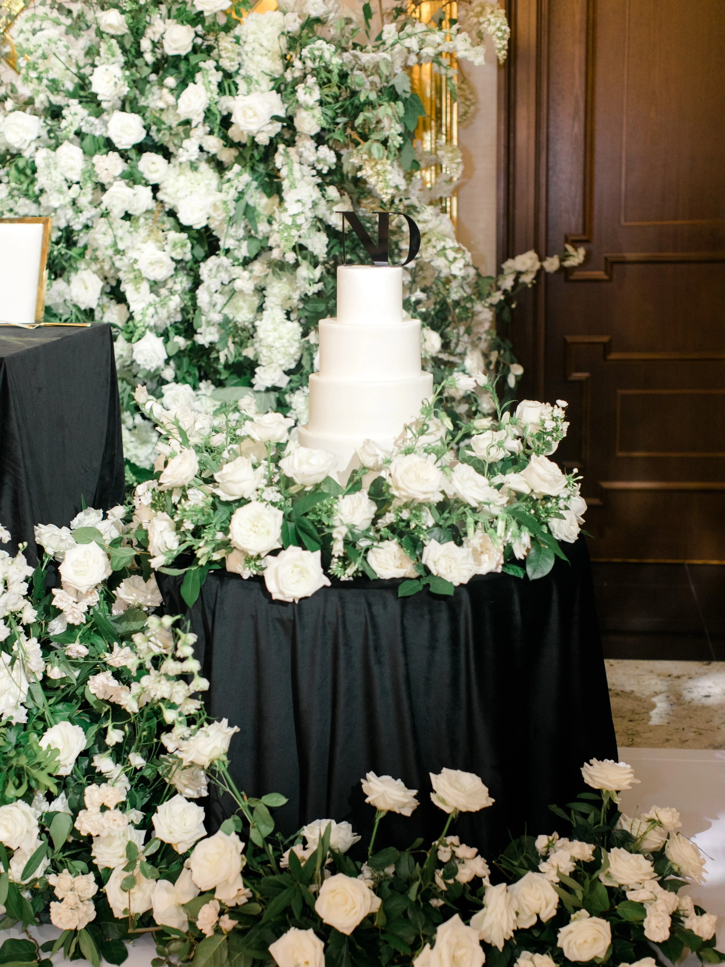 White wedding cake with three tiers, decorated with white flowers and green leaves, topped with the initials 'ND', surrounded by a floral arrangement of white roses and greenery, against a backdrop of large floral display.