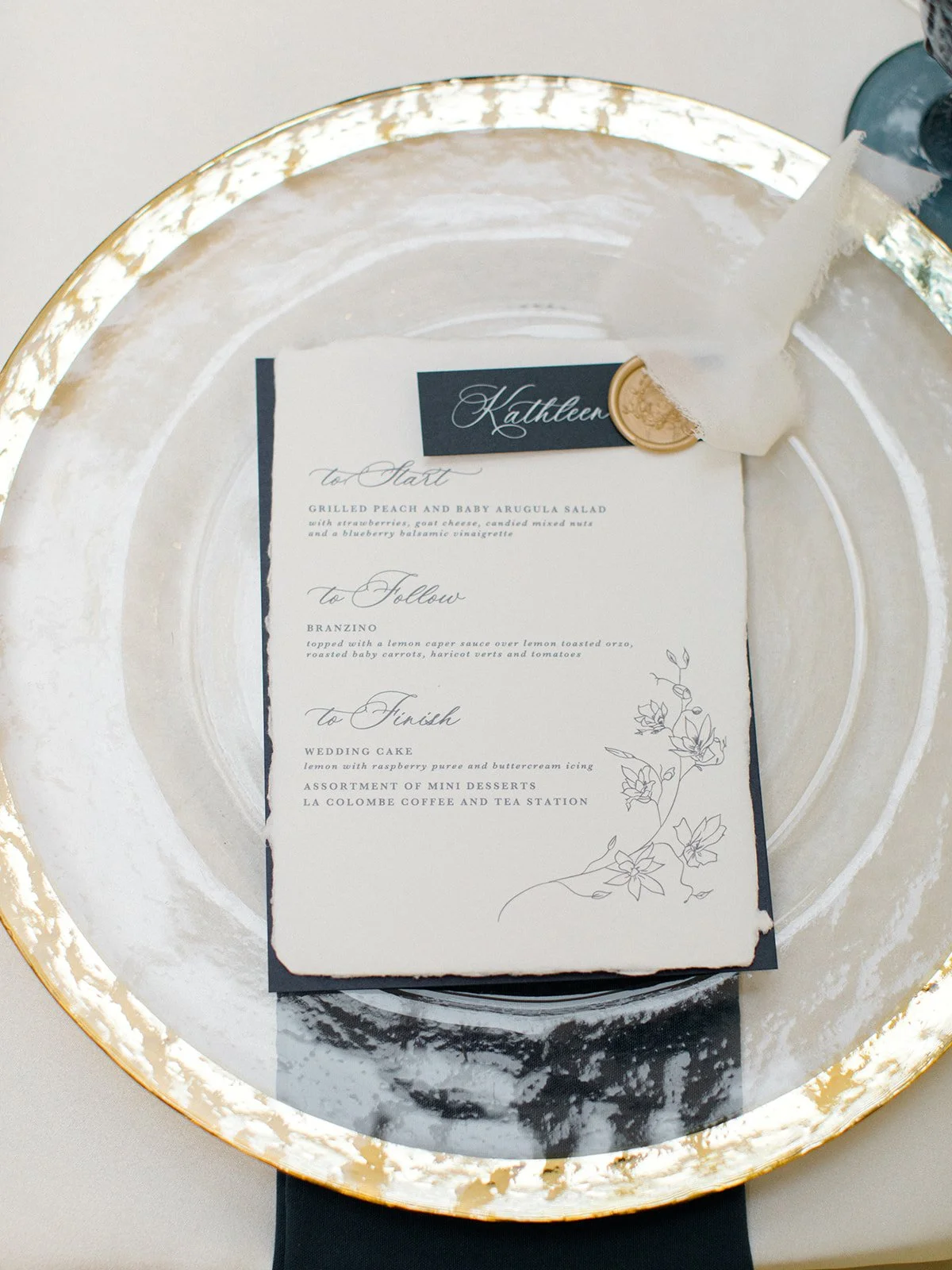 Wedding menu on a white card with floral design, placed inside a glass plate with a gold rim, on a table with a black napkin.