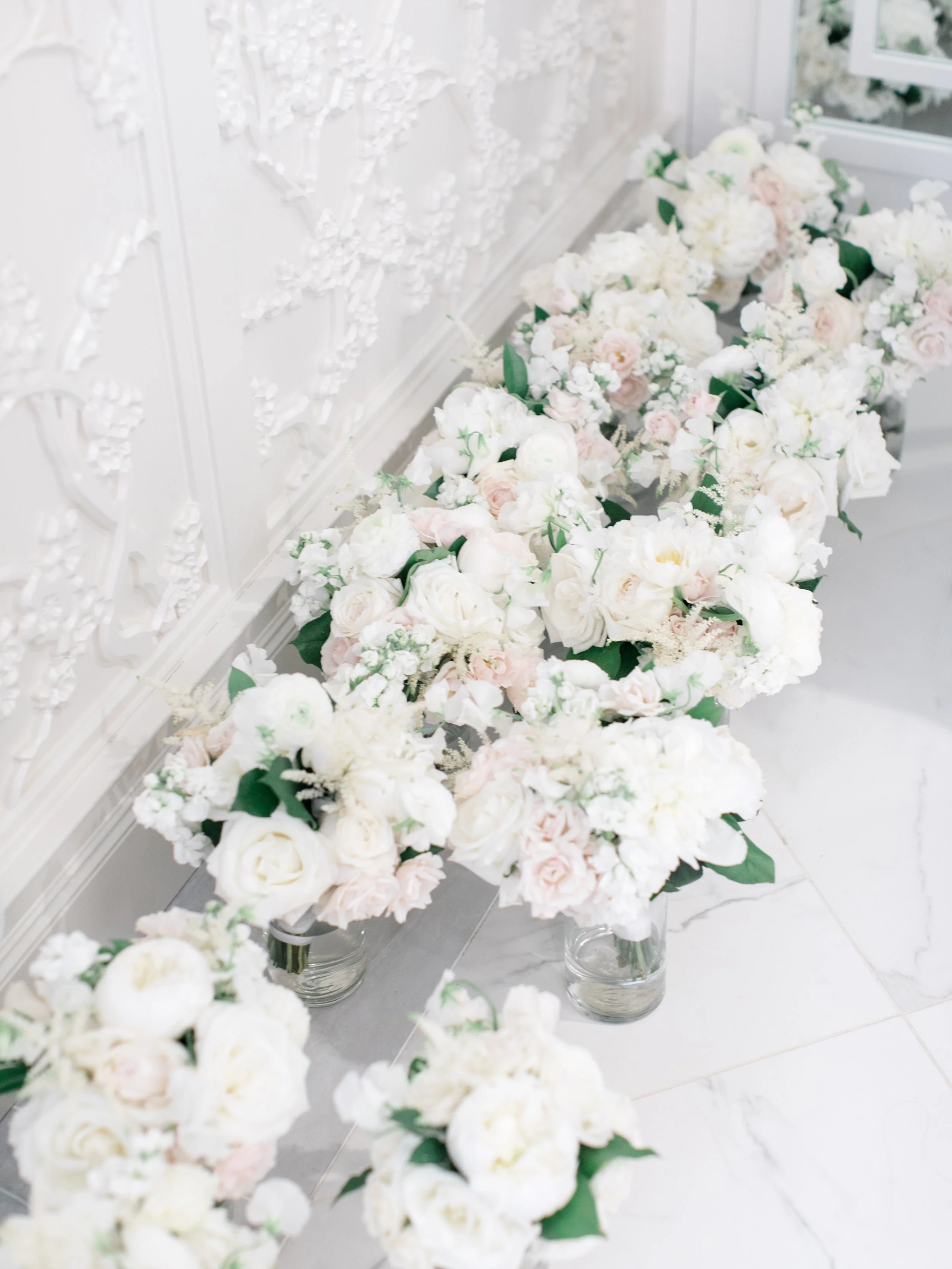 Several glass vases filled with white and pale pink flowers arranged along a white ornate wall and marble floor.