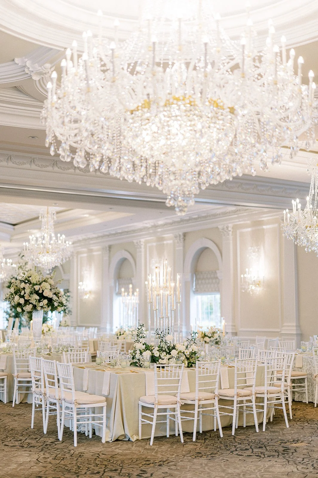 Elegant banquet hall decorated with multiple large crystal chandeliers, white chairs, and floral centerpieces featuring white flowers and greenery.