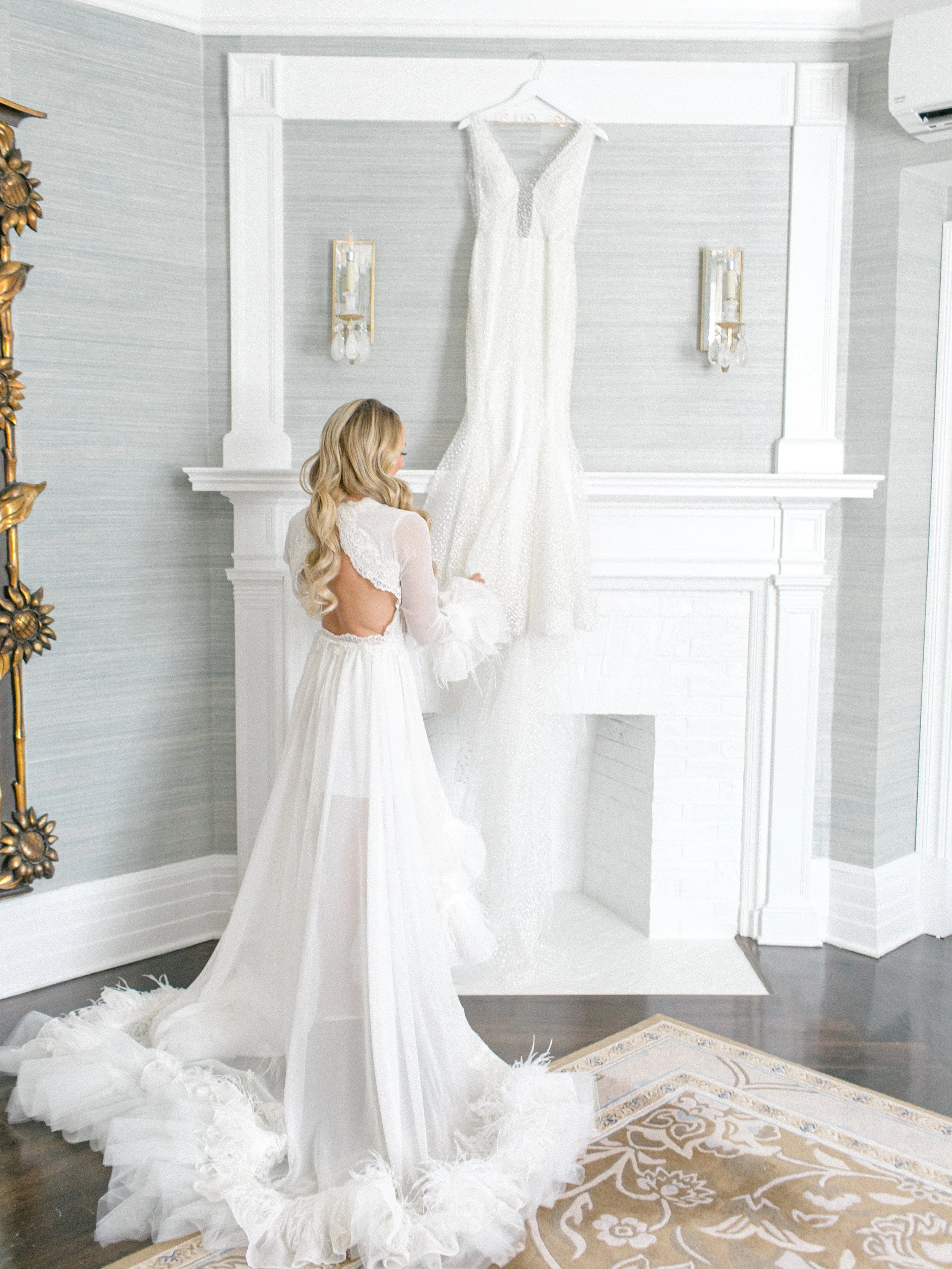 A woman in a white wedding dress holding a feathered fan, standing in front of a hanging wedding gown above a fireplace in a bright, modern room.