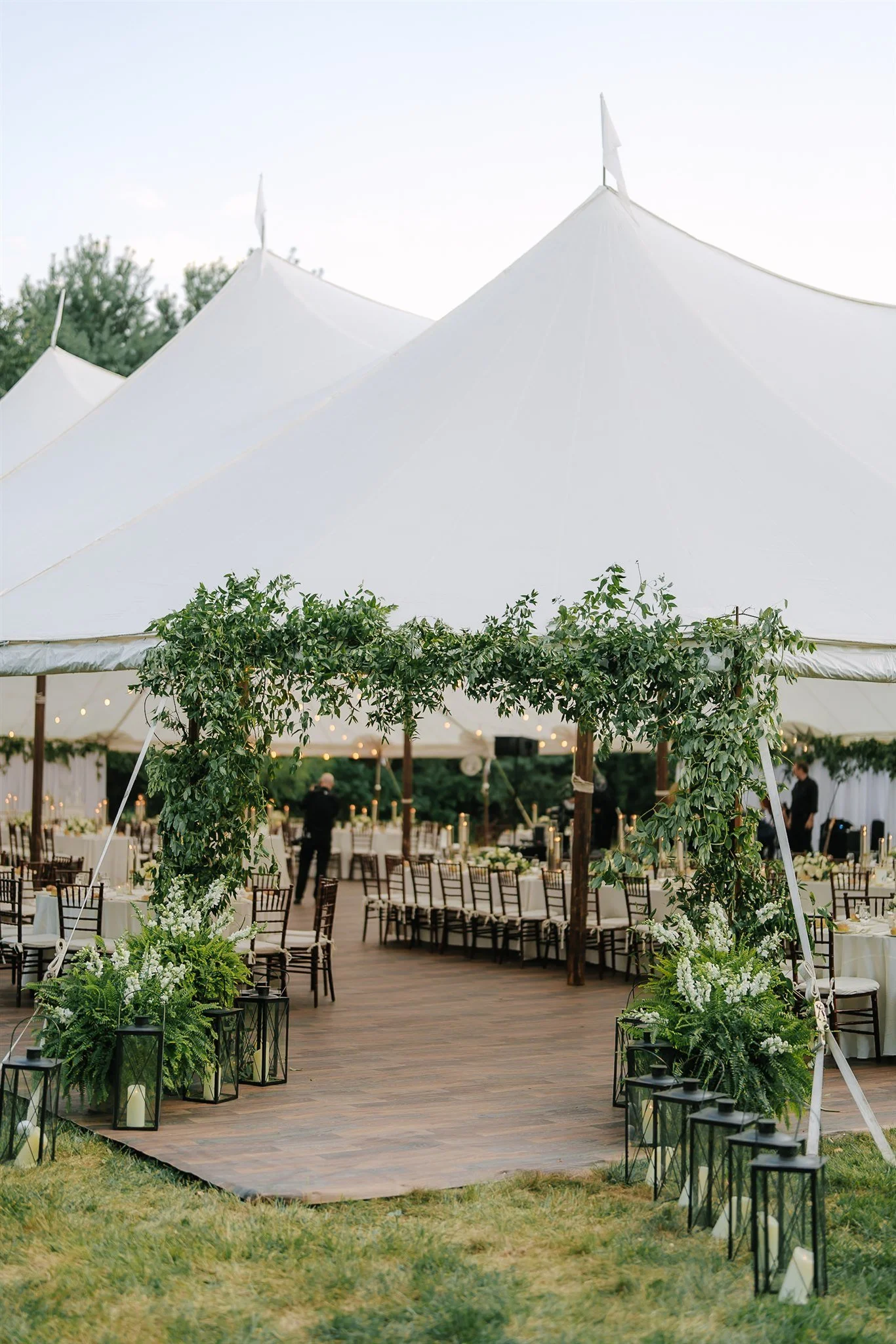 An outdoor wedding setup under a large white tent with wooden flooring, decorated with green foliage and white flowers, with tables and chairs arranged for guests.