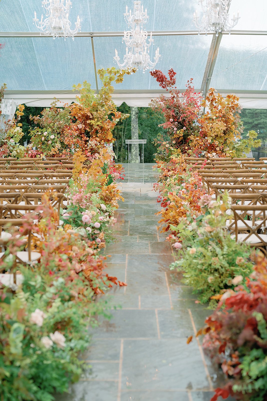 A wedding aisle decorated with colorful autumn leaves and flowers, under a clear tent with a chandelier hanging from the ceiling. A podium is at the end of the aisle.