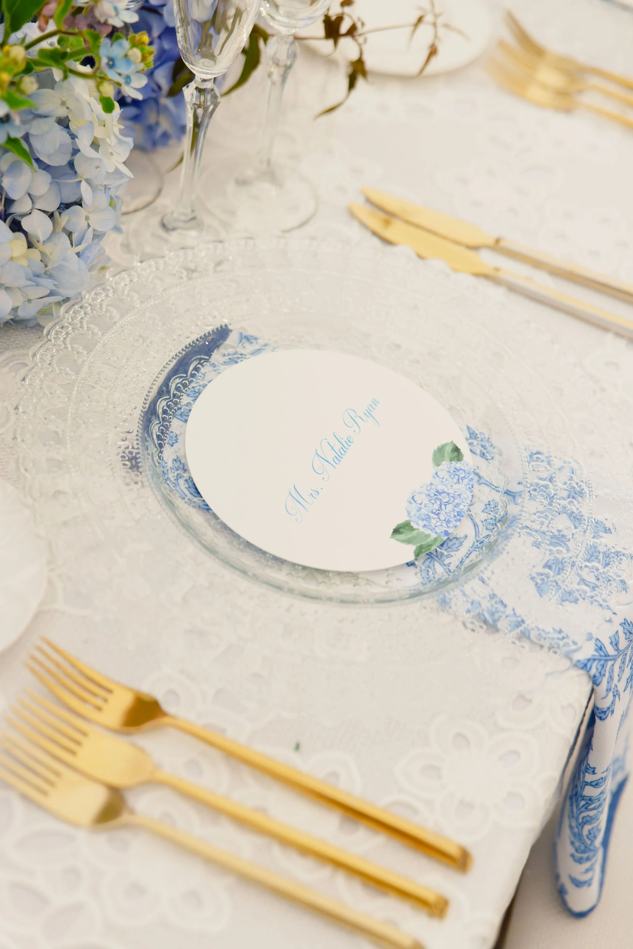 Elegant table setting with gold cutlery, a clear glass plate, a white place card with blue script reading 'Ms. Natalie Ryan,' and blue and white floral decorations.