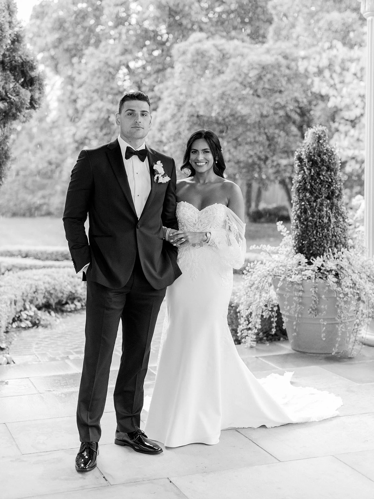A black-and-white photo of a newlywed couple standing outdoors on a stone patio, with trees and large potted plants in the background. The groom is in a black tuxedo with a bow tie, and the bride is in a strapless, off-the-shoulder white wedding gown