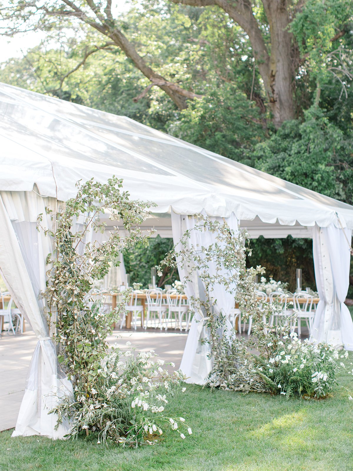 White outdoor tent decorated with greenery and white flowers, set up for an event, surrounded by lush trees and grass.